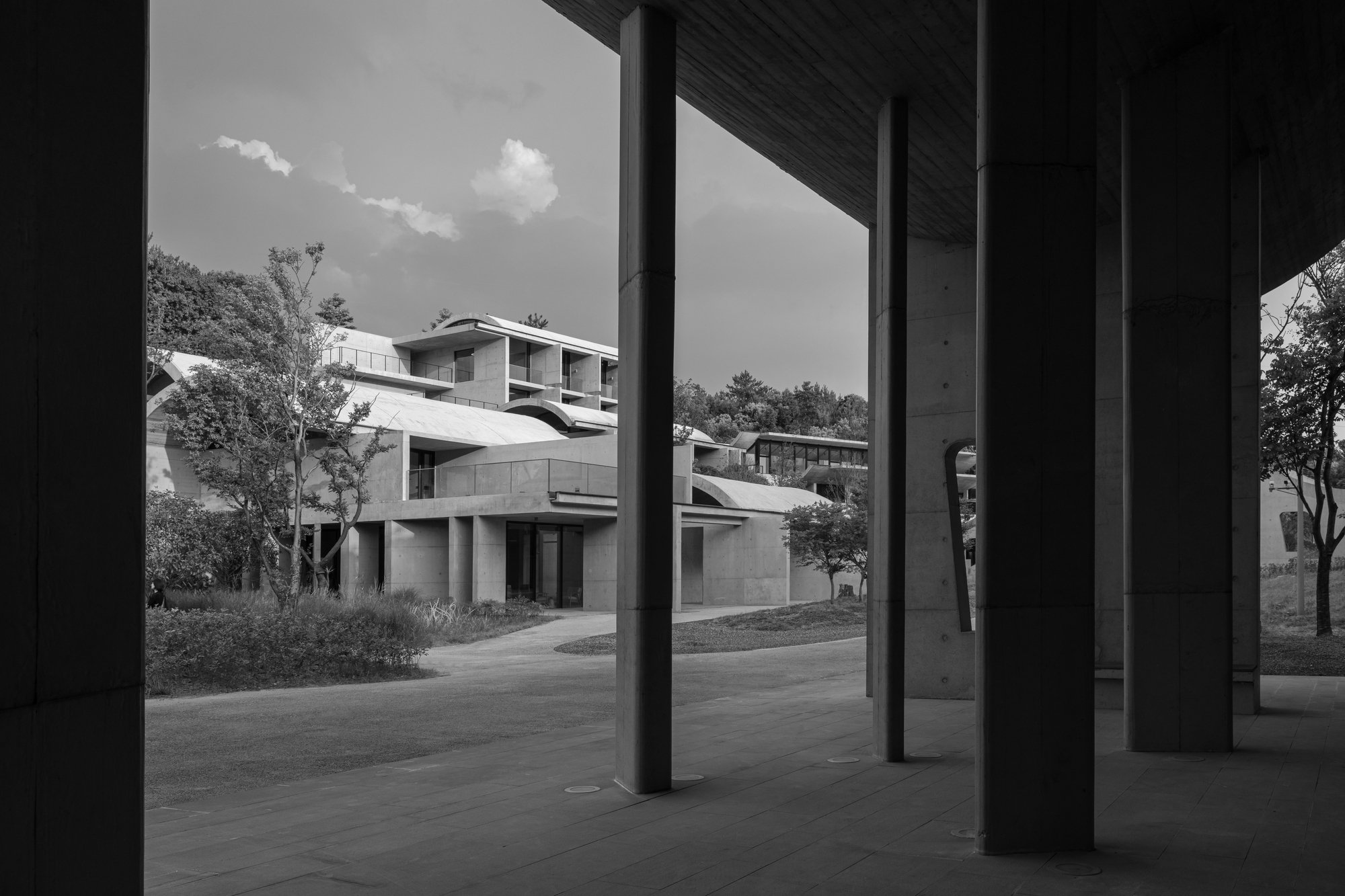 Black and white photo showing modern buildings with large windows and terraces, trees, and a partly cloudy sky viewed from a shaded patio area with concrete pillars and a tiled floor.