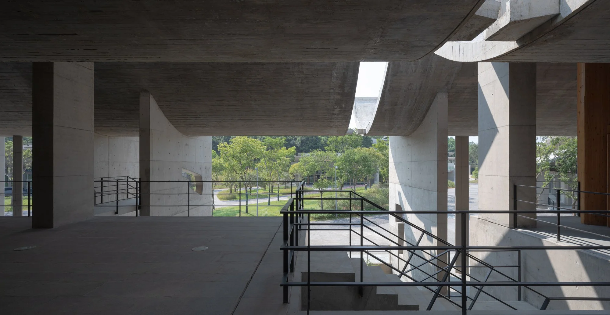 View of a modern concrete building interior with large open spaces, multiple levels, and open windows showing green trees and a park outside.