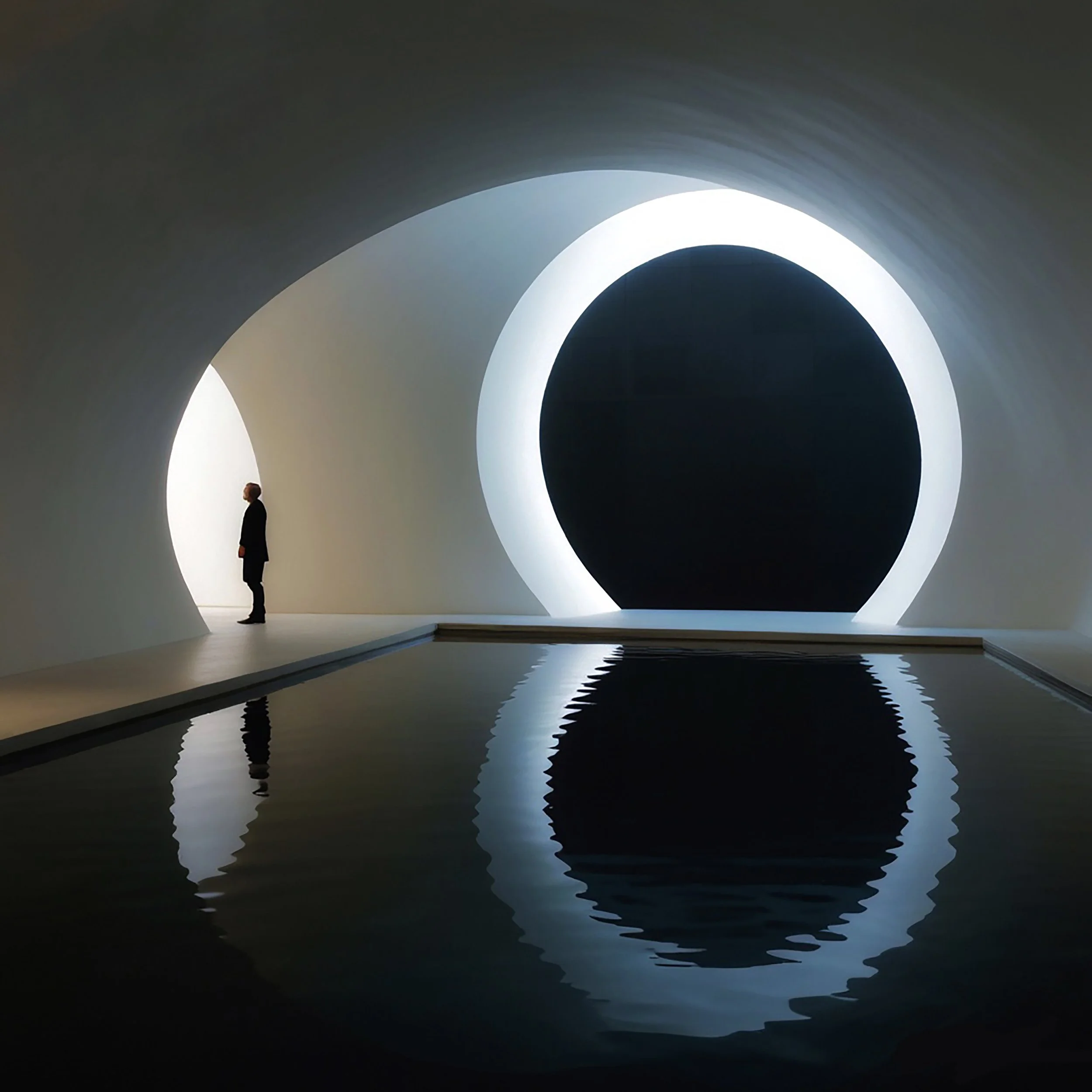 A person standing near a large, circular, illuminated structure with a reflective pool of water on the floor, creating a mirror reflection, in a dark, modern interior space.