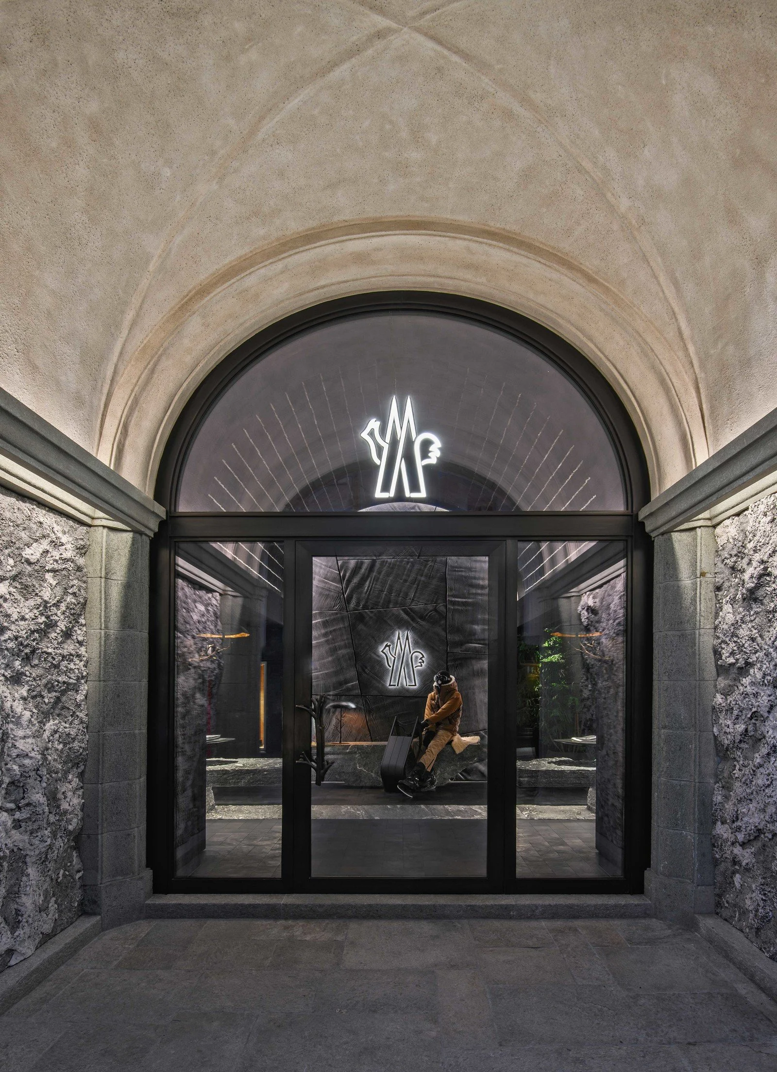 A person sitting on a bench inside a building with large glass doors and a neon sign above that features a stylized head and hands. The building has stone walls and an arched ceiling.