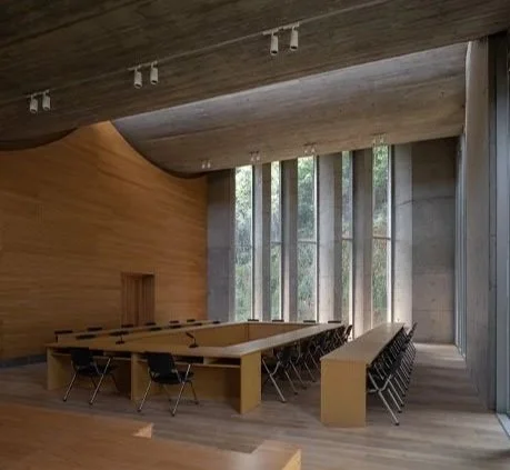 Modern conference room with a large rectangular table, black chairs, wooden panel walls, and tall vertical windows letting in natural light.