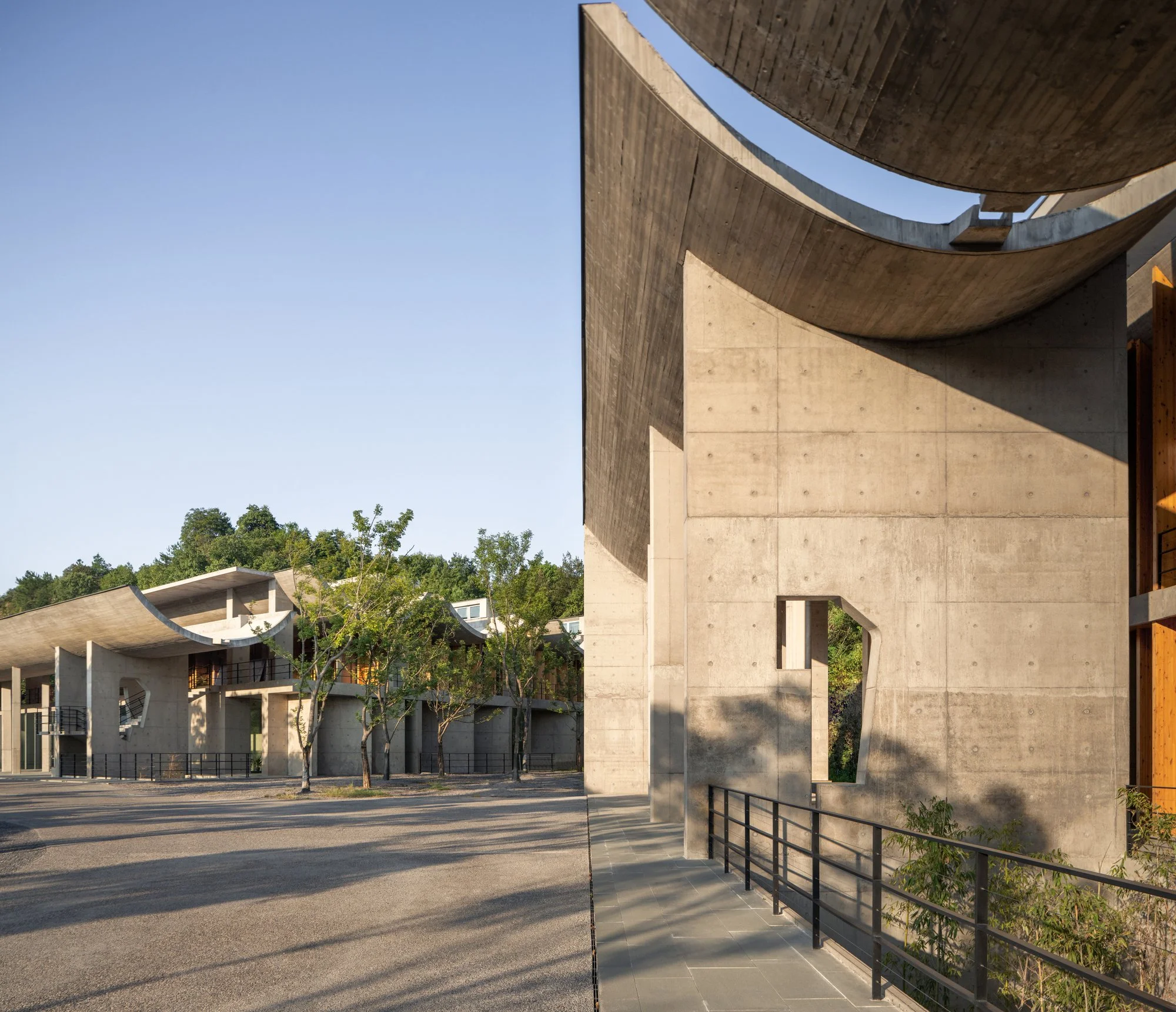 Modern architectural building with curved concrete walls and a staircase, surrounded by trees and an open parking area under a clear blue sky.
