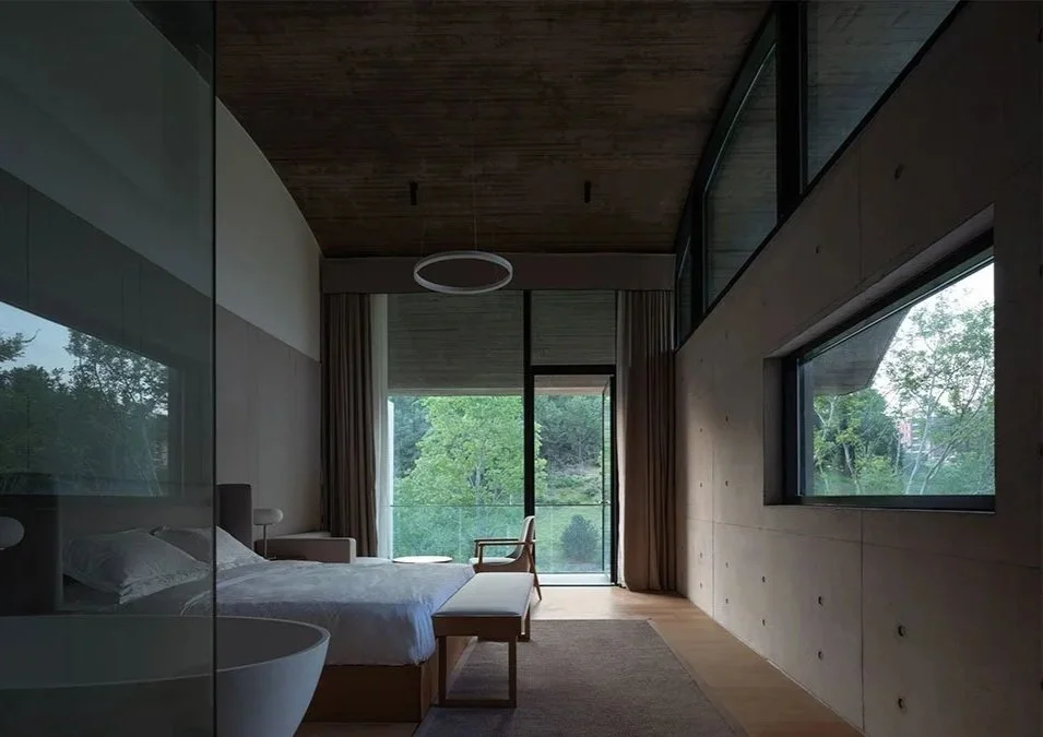 Modern bedroom with large glass windows showing green trees outside, minimalist furniture, and a wooden ceiling.