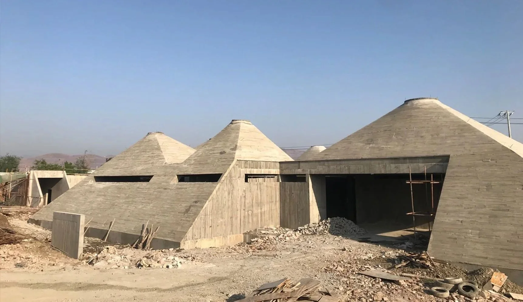 Construction site with building structures featuring cone-shaped roofs made of concrete blocks, with some metal scaffolding and construction materials around.