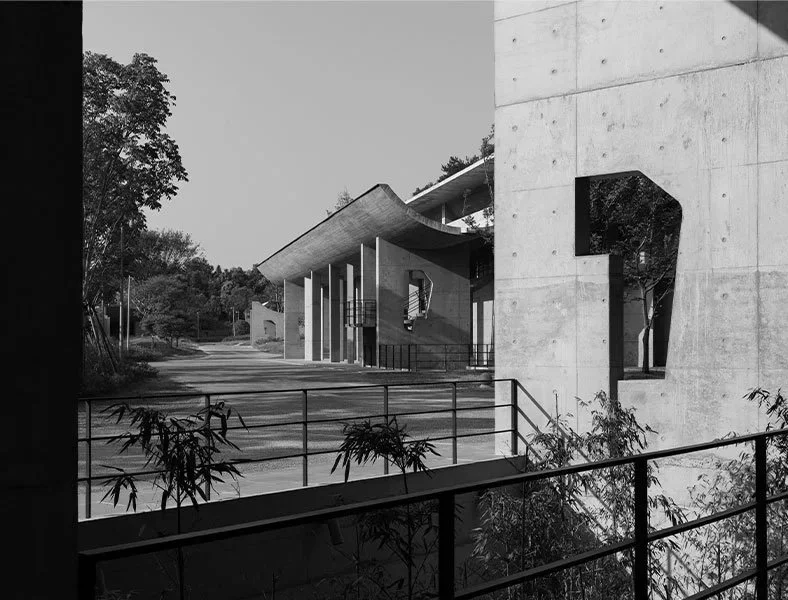 A modern architectural building with sharp, angular roof and large openings, surrounded by trees and a metal railing in black and white.