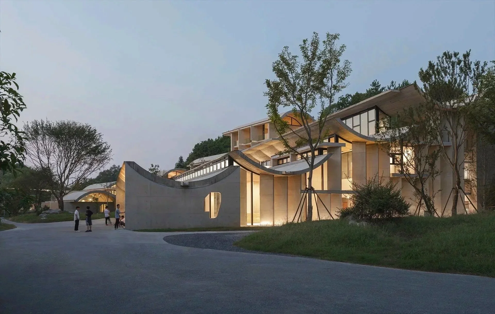 Modern architectural building with curved rooflines, large glass windows, and surrounded by trees at dusk, with a group of people standing nearby.