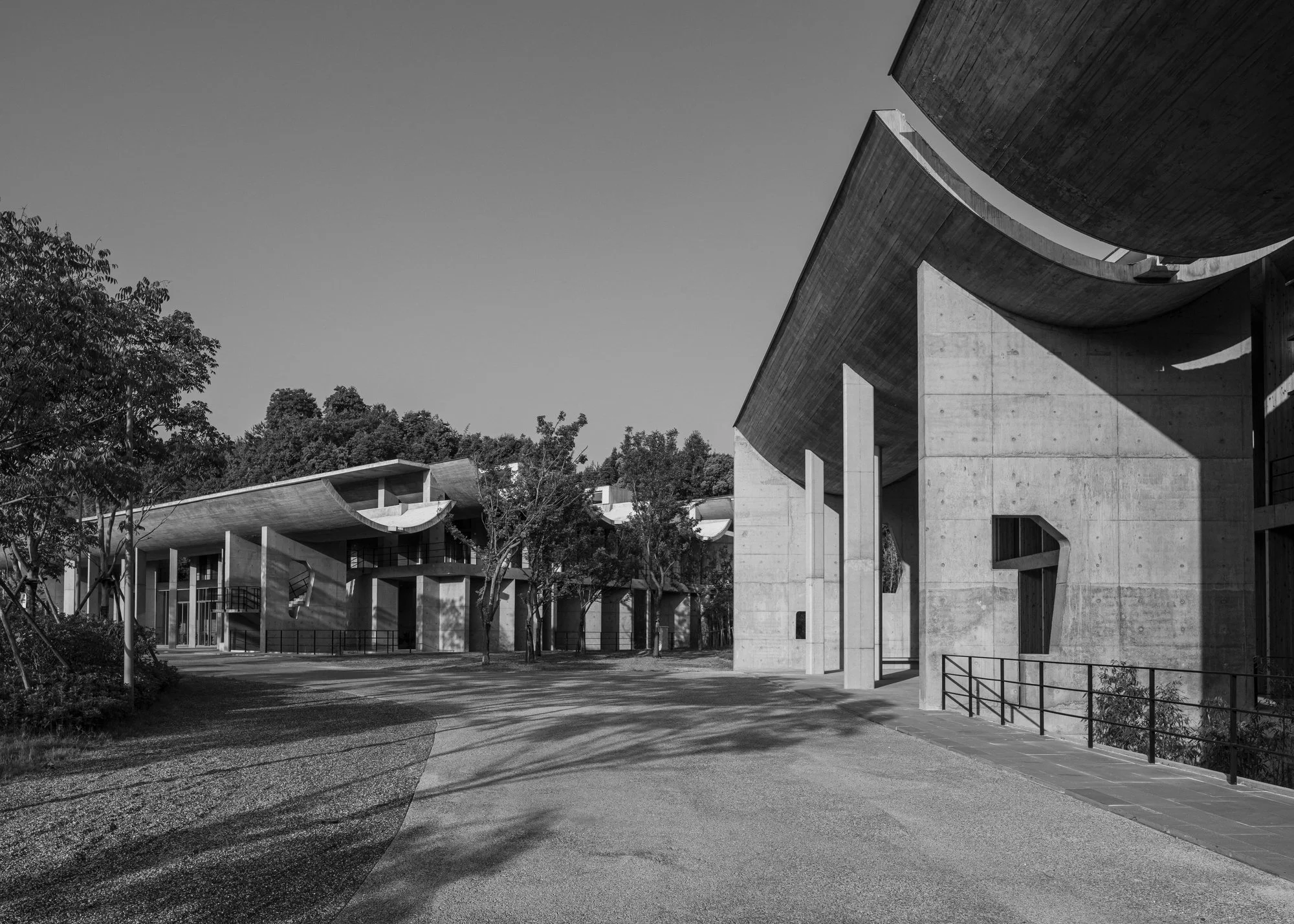 Modern concrete architectural buildings with curved roofs, surrounded by trees and a paved walkway under a clear sky.