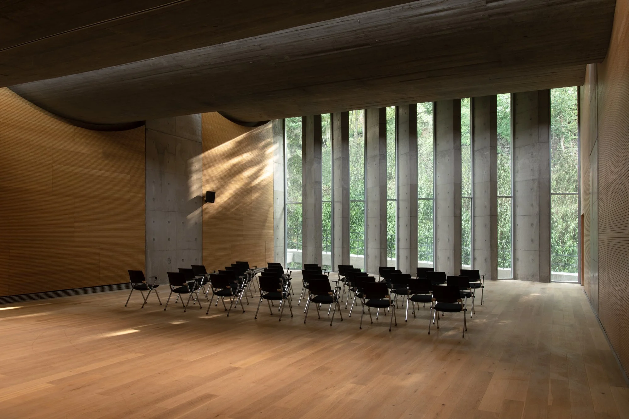 Interior of a modern conference or lecture hall with large windows, wooden flooring and walls, and black chairs arranged in rows.