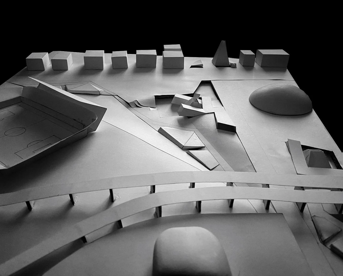 Architectural model of a modern building complex with a sports field, various geometric structures, and curved walkways, shown in black and white.
