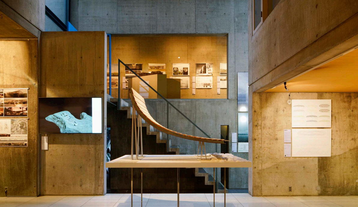 Museum interior with concrete walls, displaying photographs, diagrams, and a boat model on a table, with stairs and a monitor showing an ancient artifact.