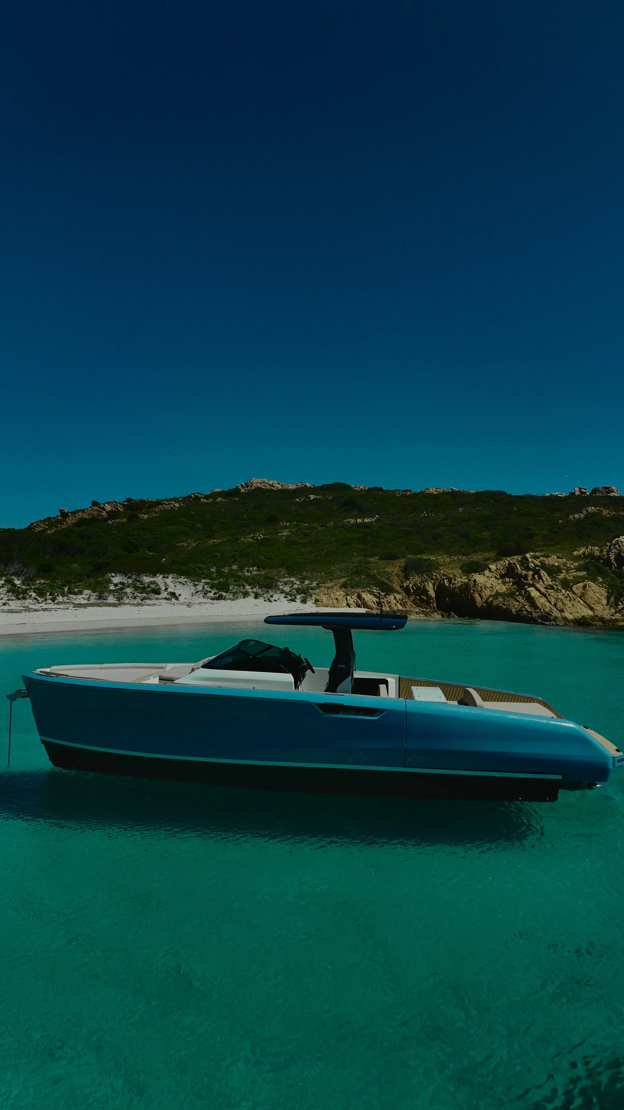 Barca a motore bianca ormeggiata in acque tranquille di un mare turchese, con una collina verde e rocciosa sullo sfondo sotto un cielo azzurro senza nuvole.