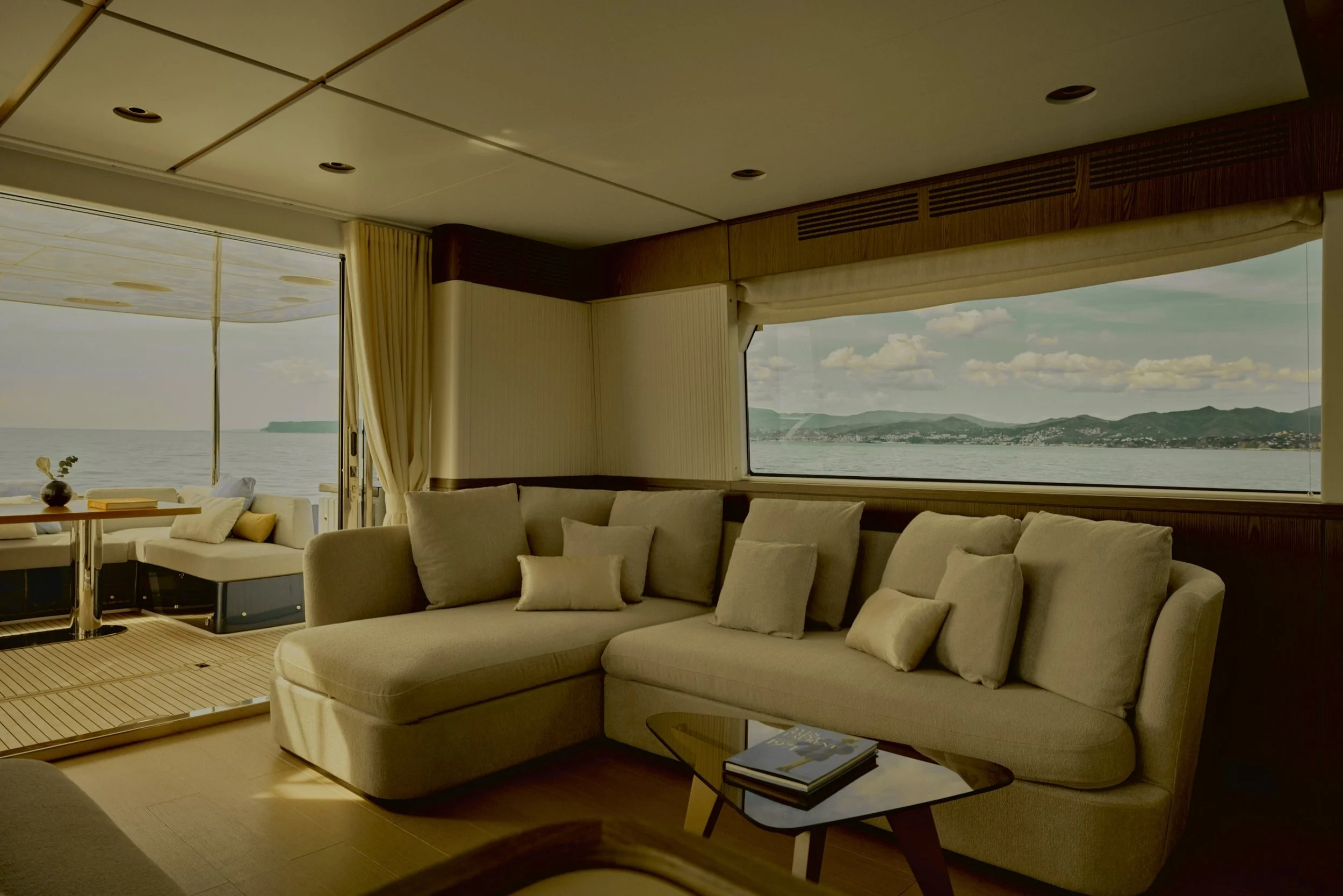 Interno di una stanza con grande finestra che mostra il mare e le colline, arredamento elegante con divani bianchi e tavolino con libri.