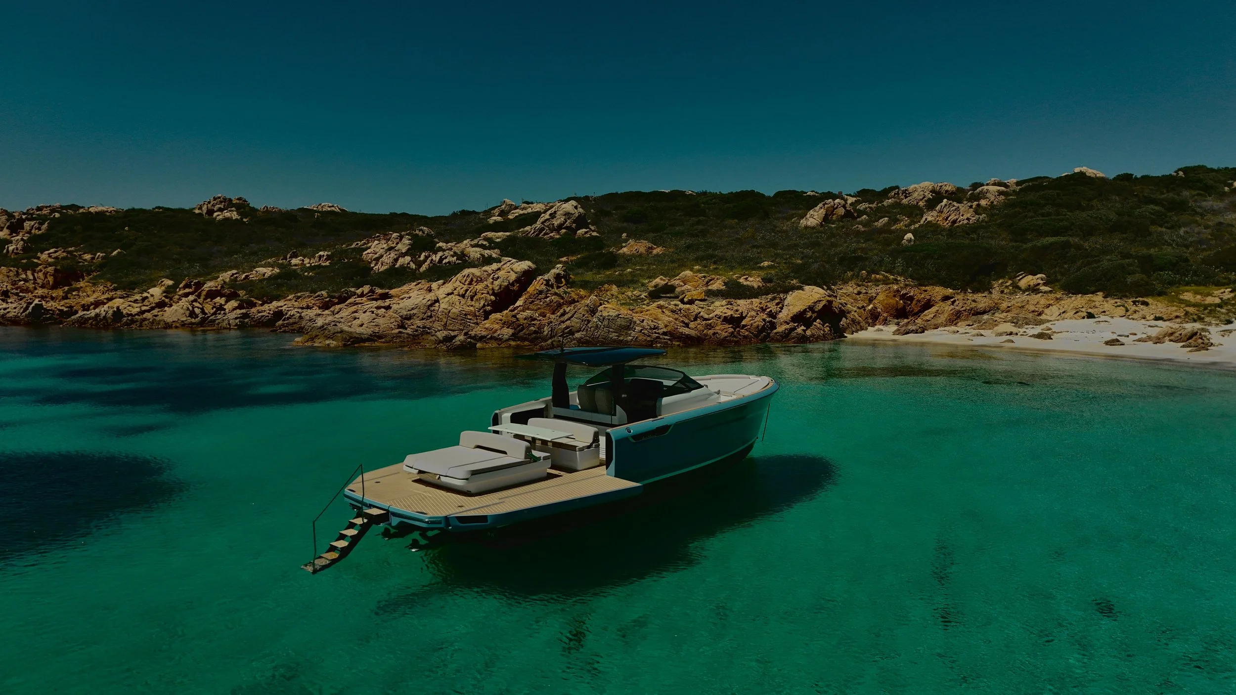Una barca a motore ancorata in un acque cristalline vicino a una spiaggia con scogli e vegetazione verde, sotto un cielo azzurro senza nuvole.