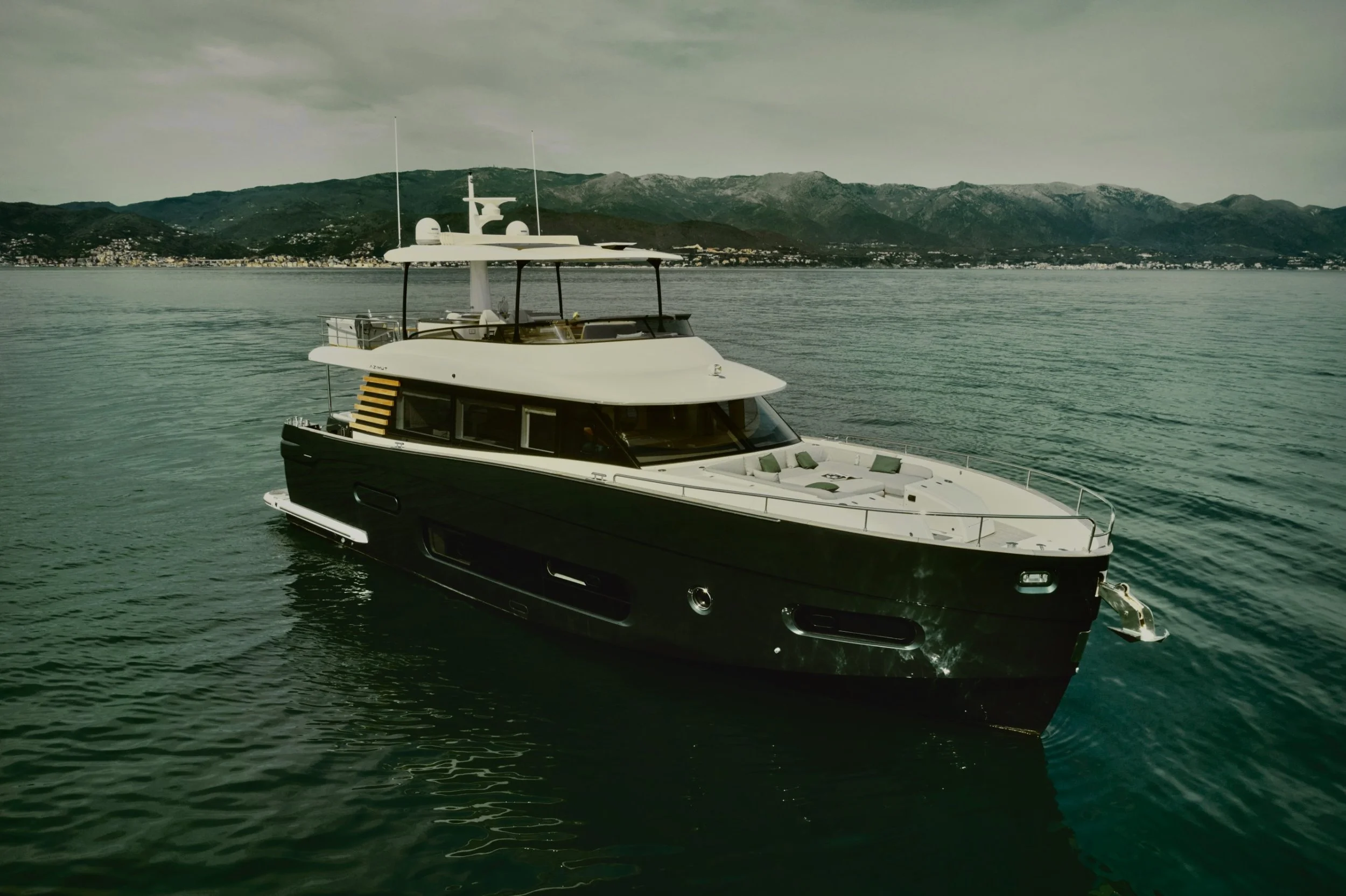 Yacht bianca ormeggiata in mare con montagne e città sullo sfondo, cielo nuvoloso.
