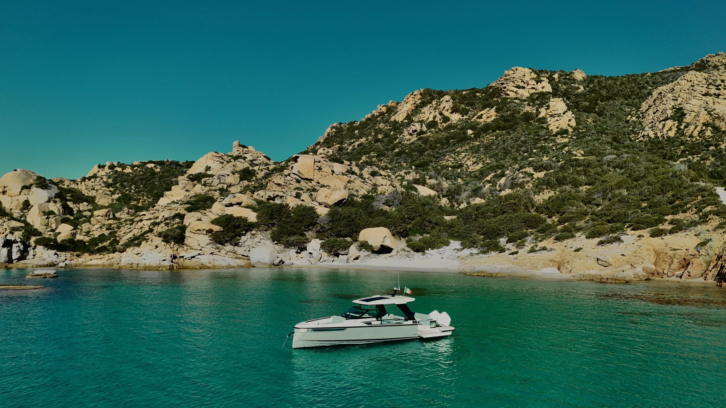 Una barca bianca si trova in acqua vicino a una costa rocciosa con vegetazione verde e alte colline o montagne sotto un cielo azzurro.