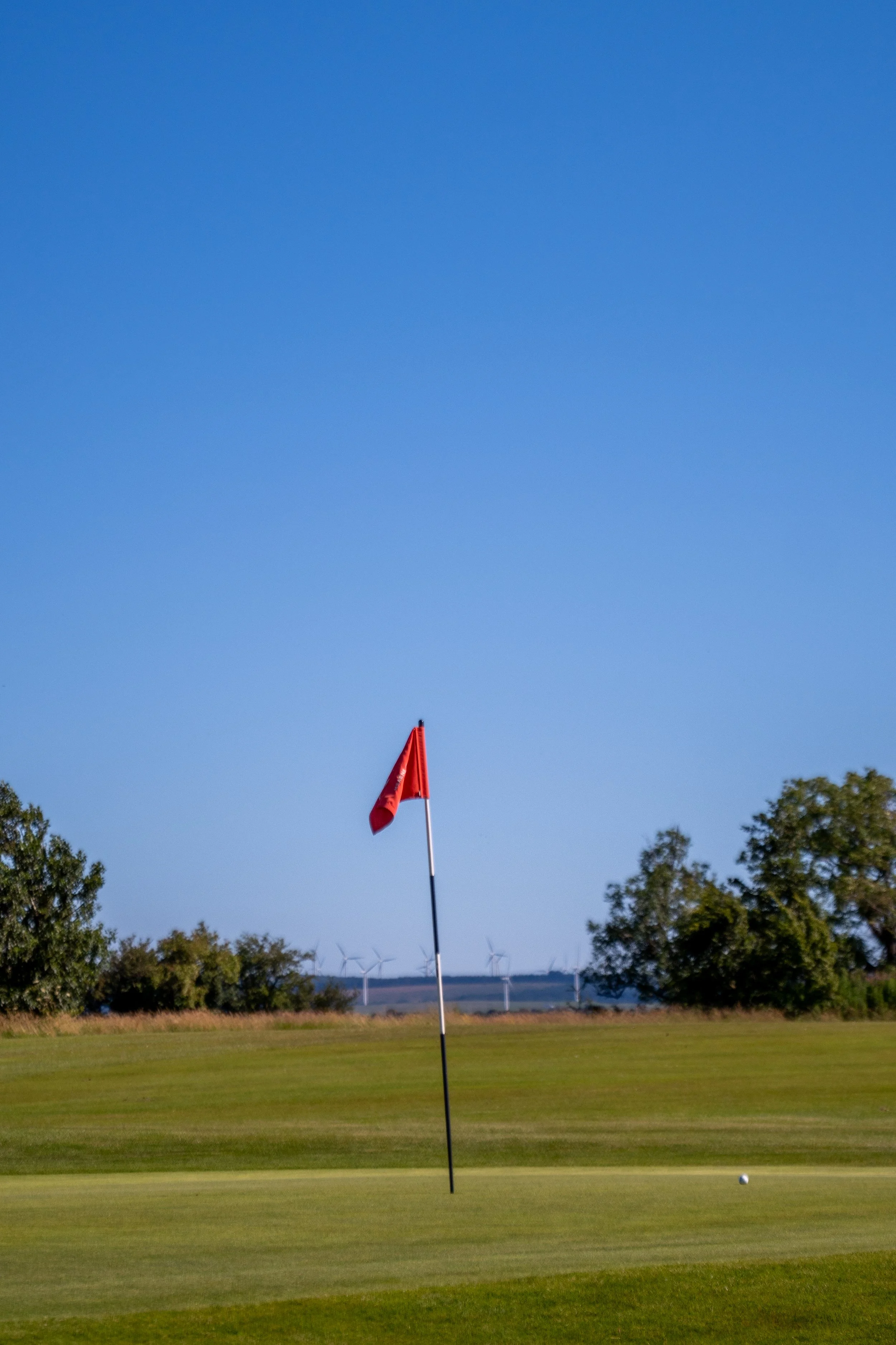 A golf course with a red flag on a green, a golf ball near the hole, trees on the horizon, and wind turbines in the distance under a blue sky.