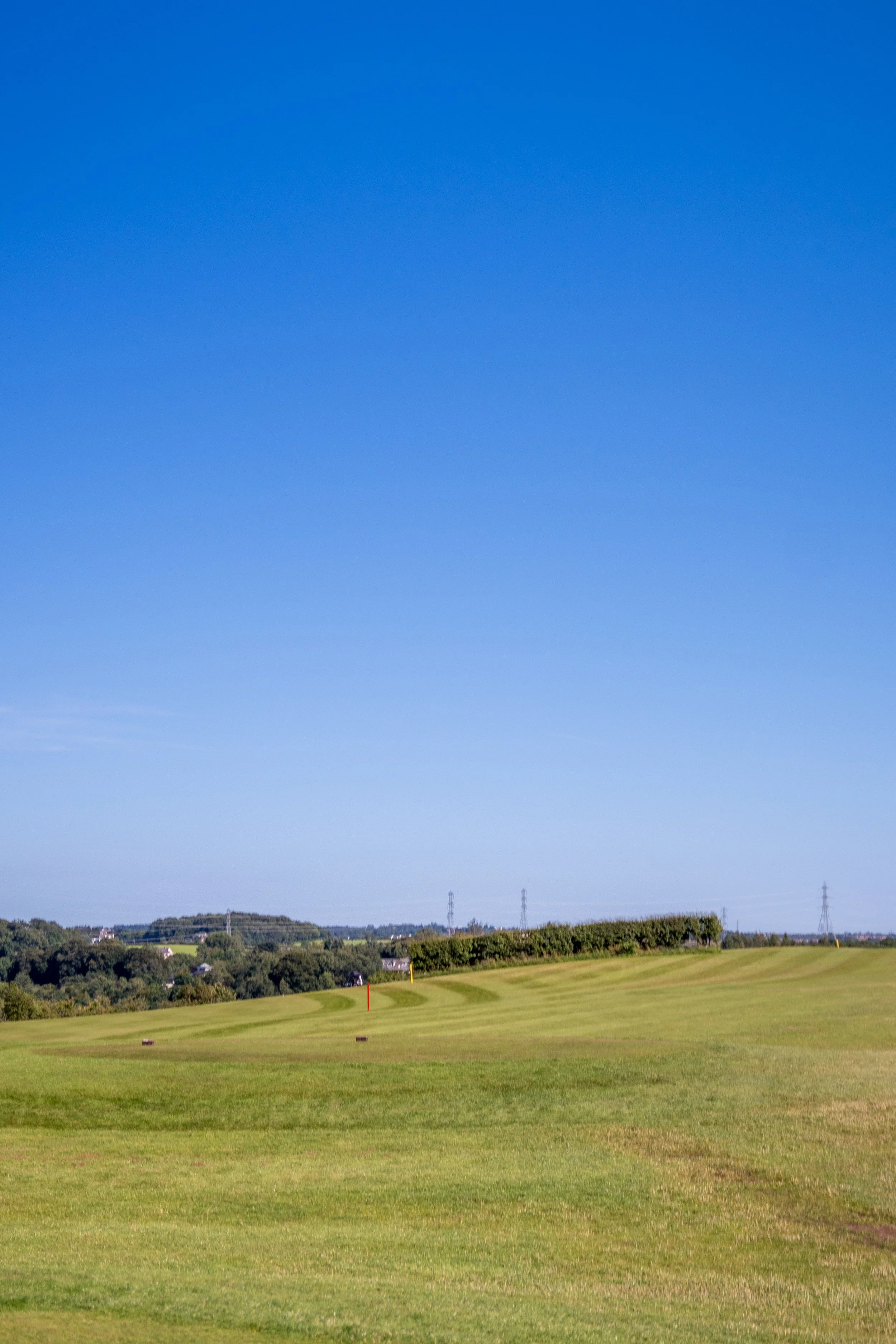 A wide view of a golf course with green grass, a few trees, and some distant hills with power lines and towers under a clear blue sky.