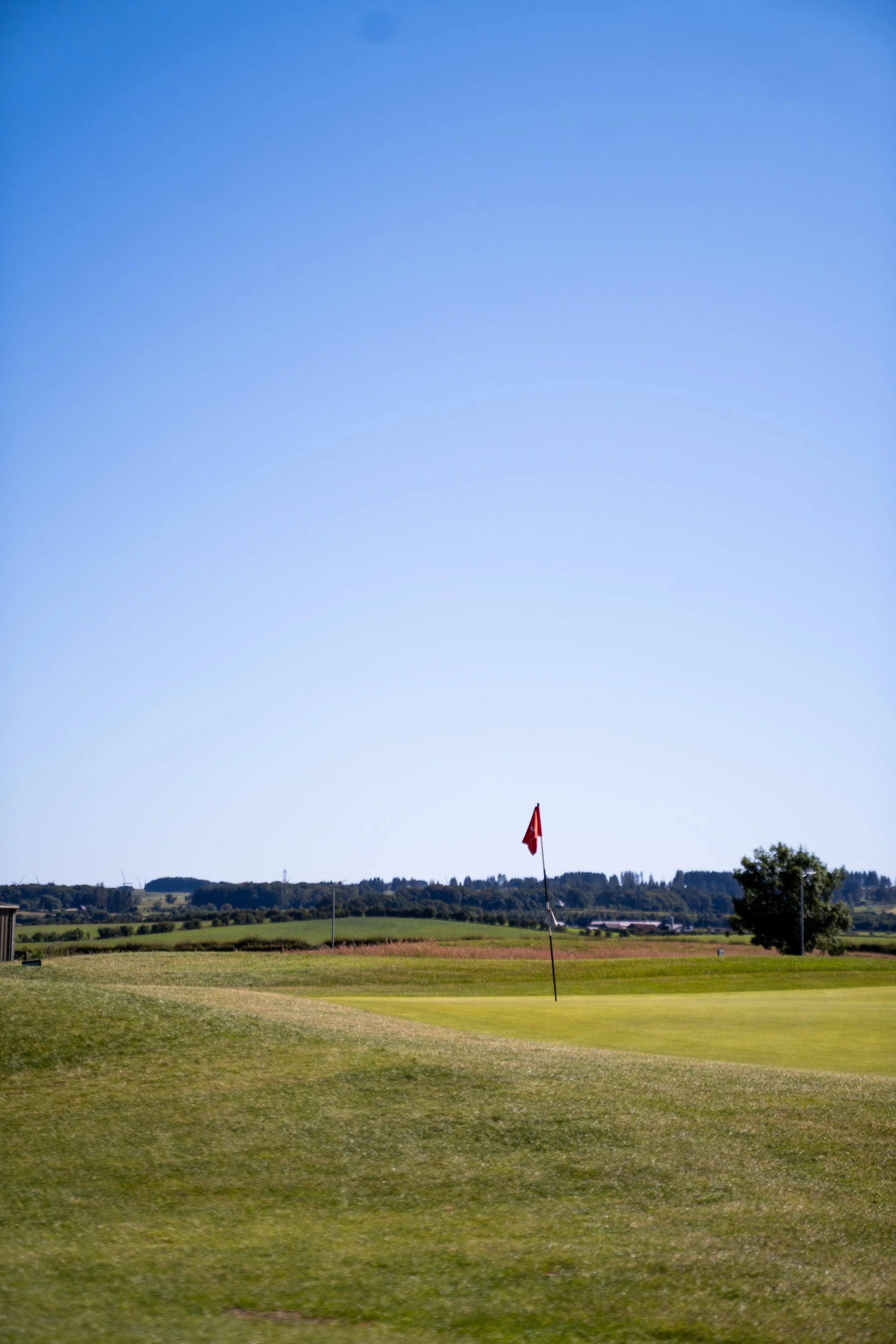 A golf course with a flag on the green and rolling hills in the background under a blue sky.