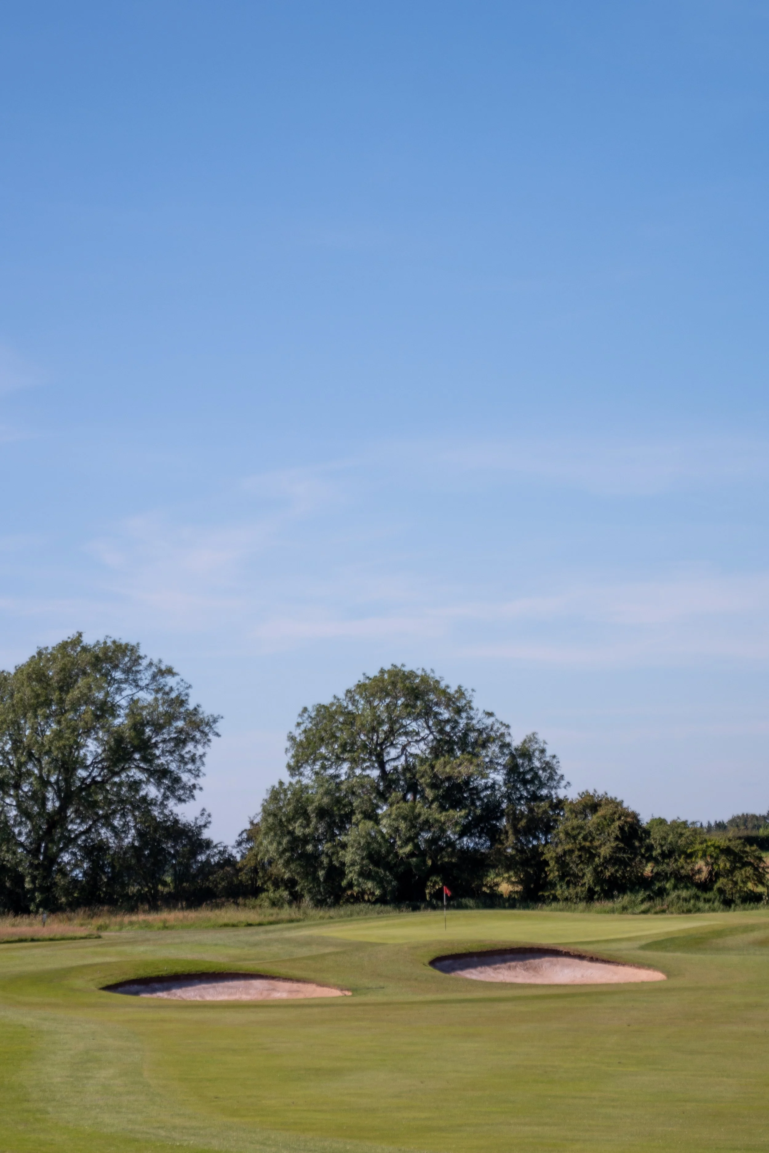 A golf course with a green, sand bunkers, a flagstick, and trees in the background under a clear blue sky.