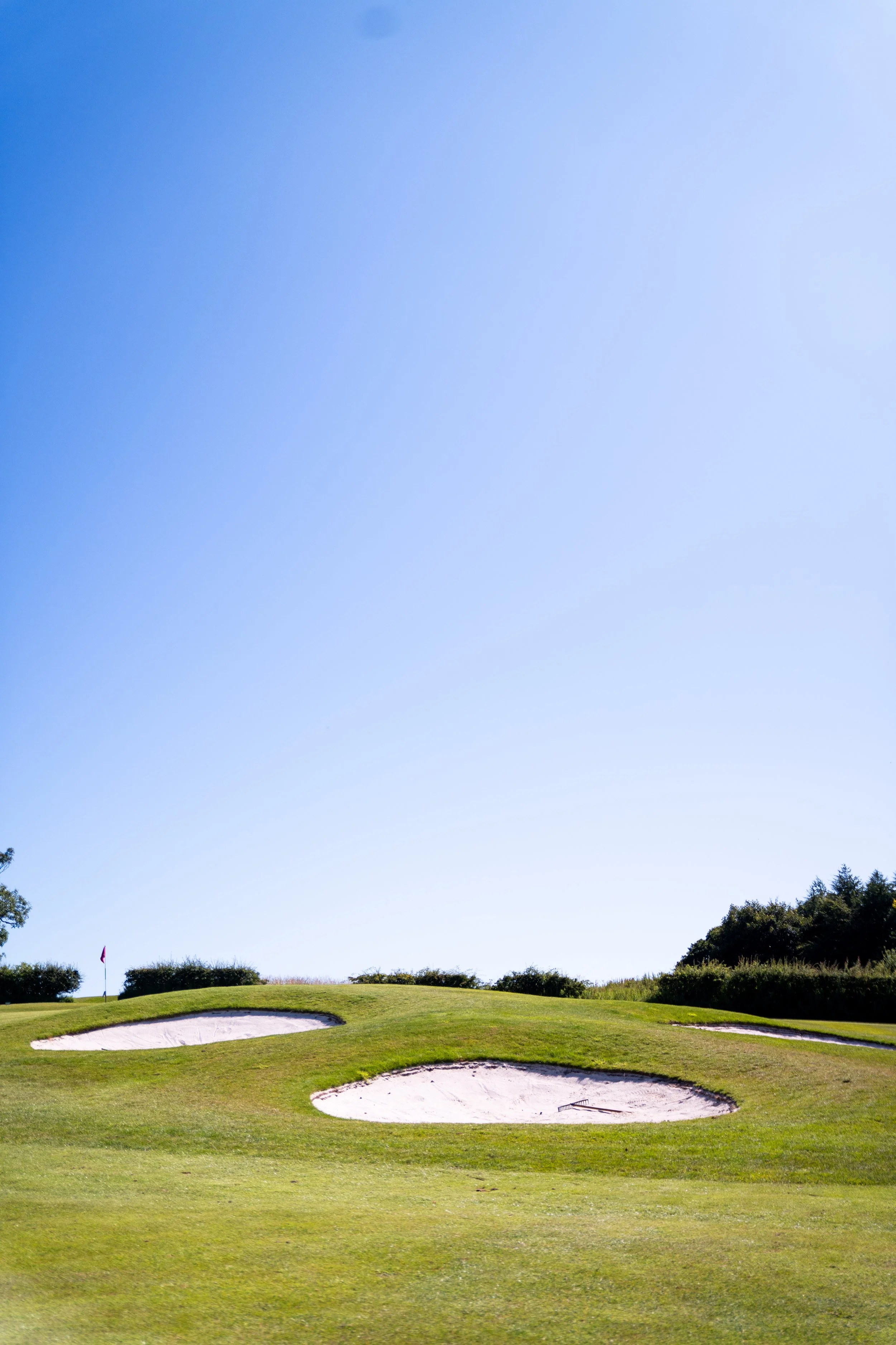 A golf course with sand bunkers, green grass, and a flagstick on the green, under a clear blue sky with some trees in the background.