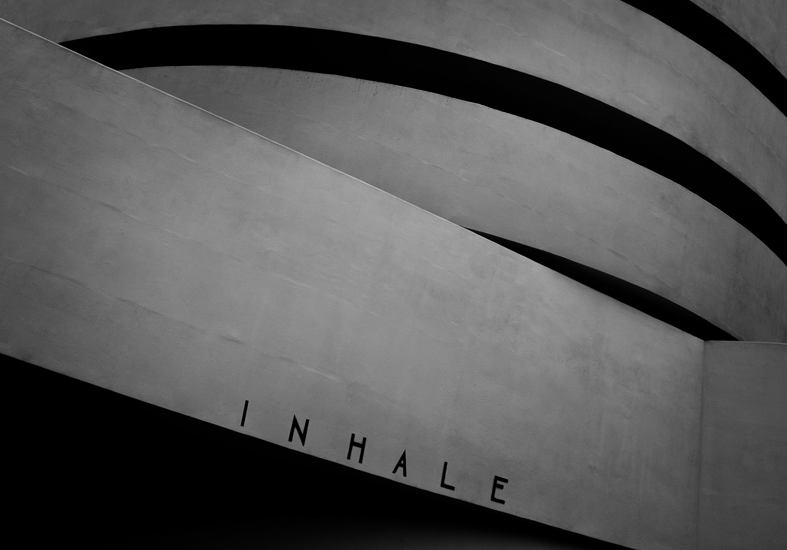 Close-up of a modern architectural structure with curved concrete slabs and the word 'INHALE' written in black letters on a concrete surface.