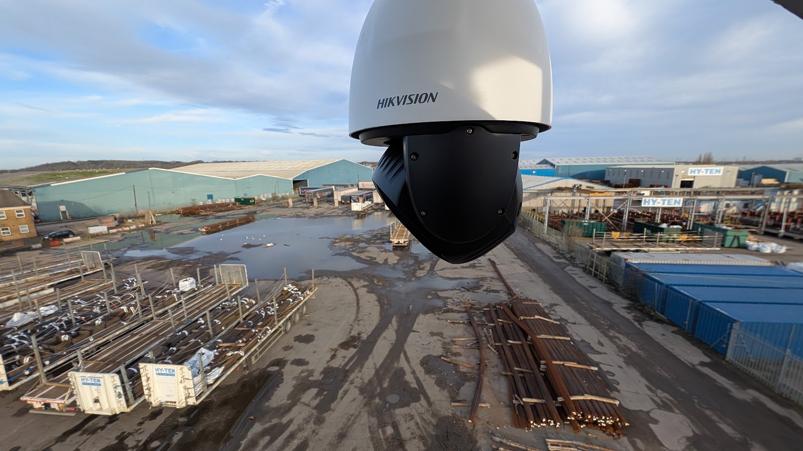 Close-up of a Hikvision surveillance camera mounted outdoors, overlooking an industrial yard with scattered metal rails, containers, and warehouses under a partly cloudy sky.