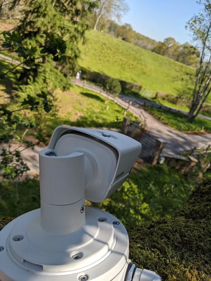 A surveillance camera mounted on a pole overlooks a green scenic park with a pathway and small bridge in the background.