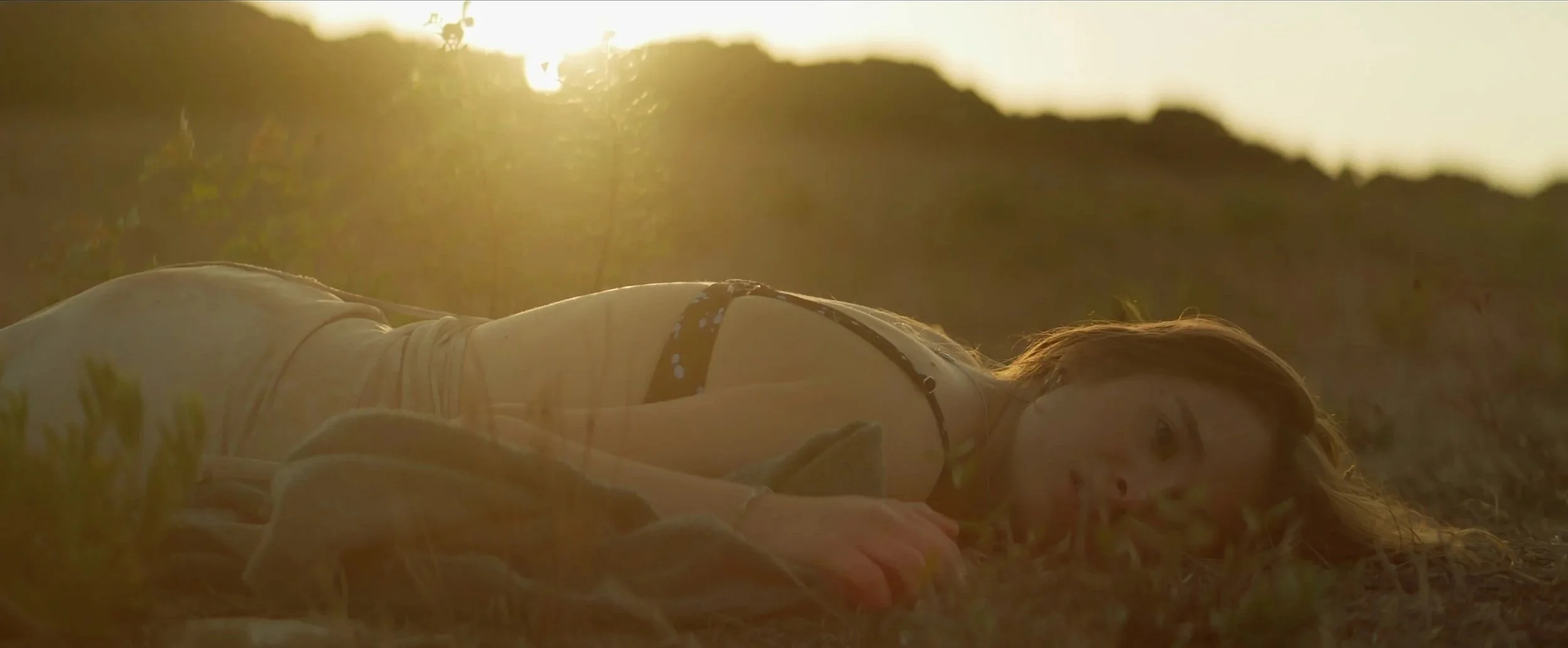 Una donna sdraiata sull'erba al tramonto, con i capelli castani e un vestito a spalline, che guarda verso la fotocamera con uno sfondo naturale e un cielo dorato.