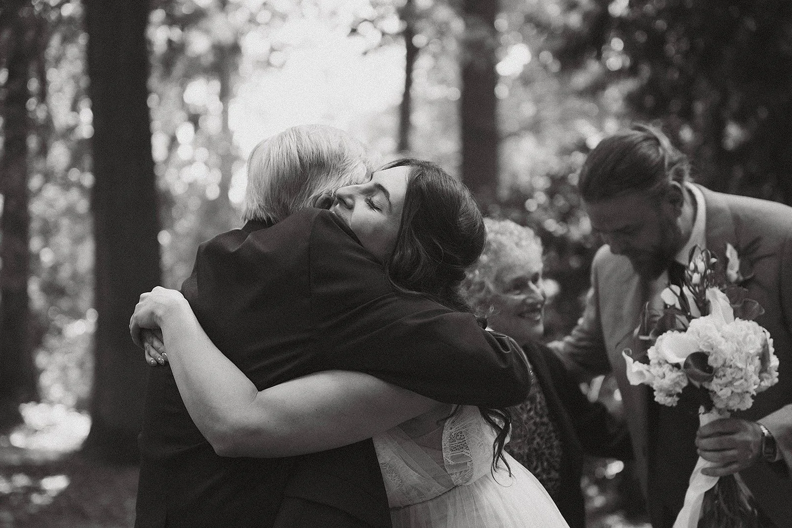 A woman in a wedding dress embraces an older man in a forest, as a man holding a bouquet of flowers smiles nearby, with a woman in glasses watching.