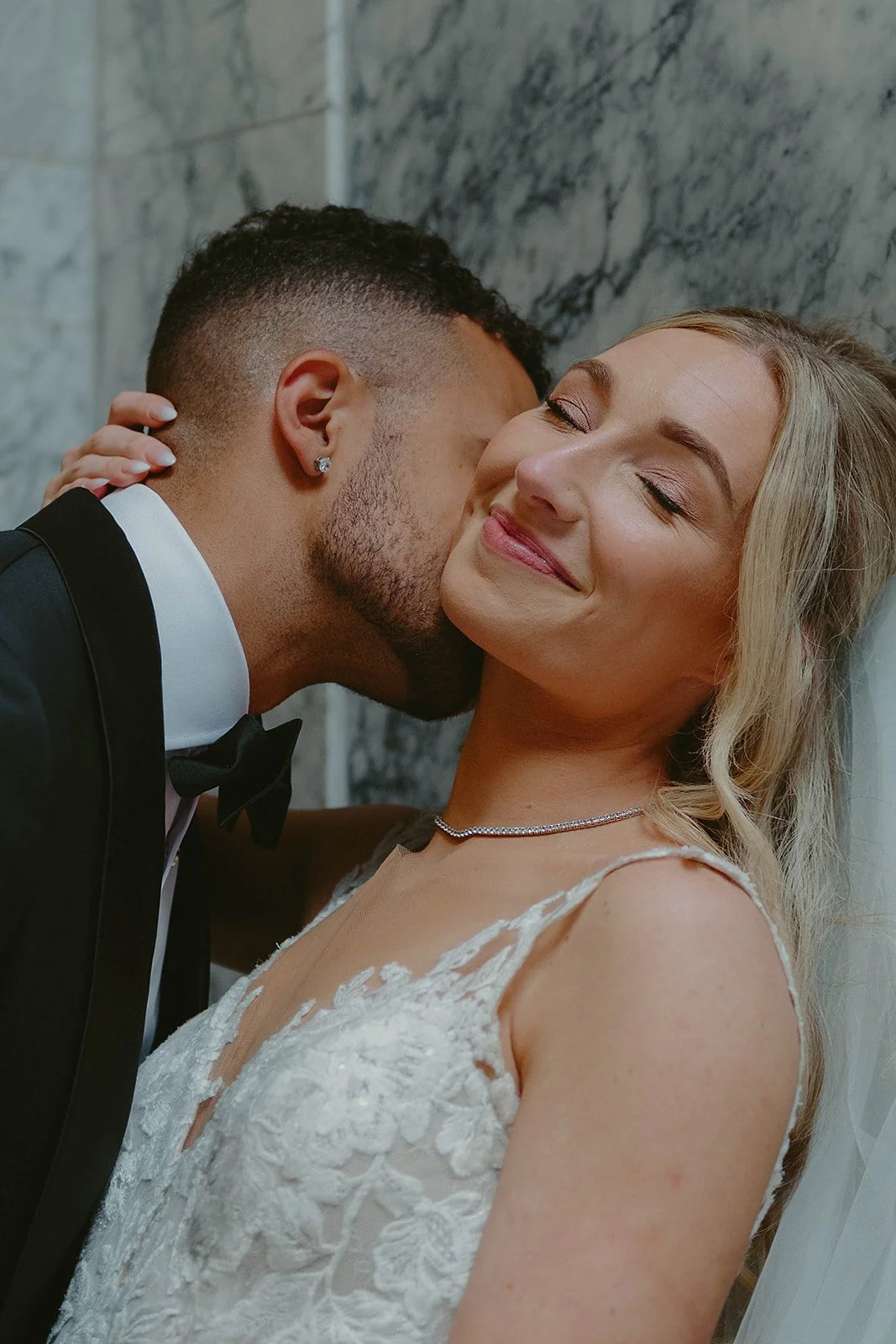A groom kissing a bride on the cheek during their wedding, with a marble wall background.