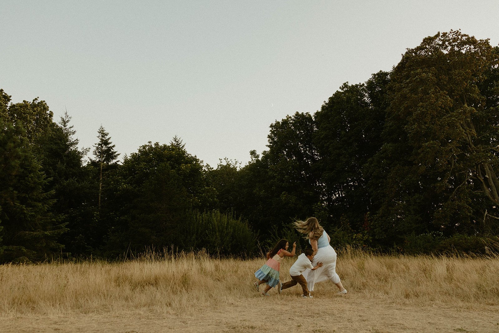 A woman and two children are playing and running in a grassy field with trees in the background under a cloudy sky.