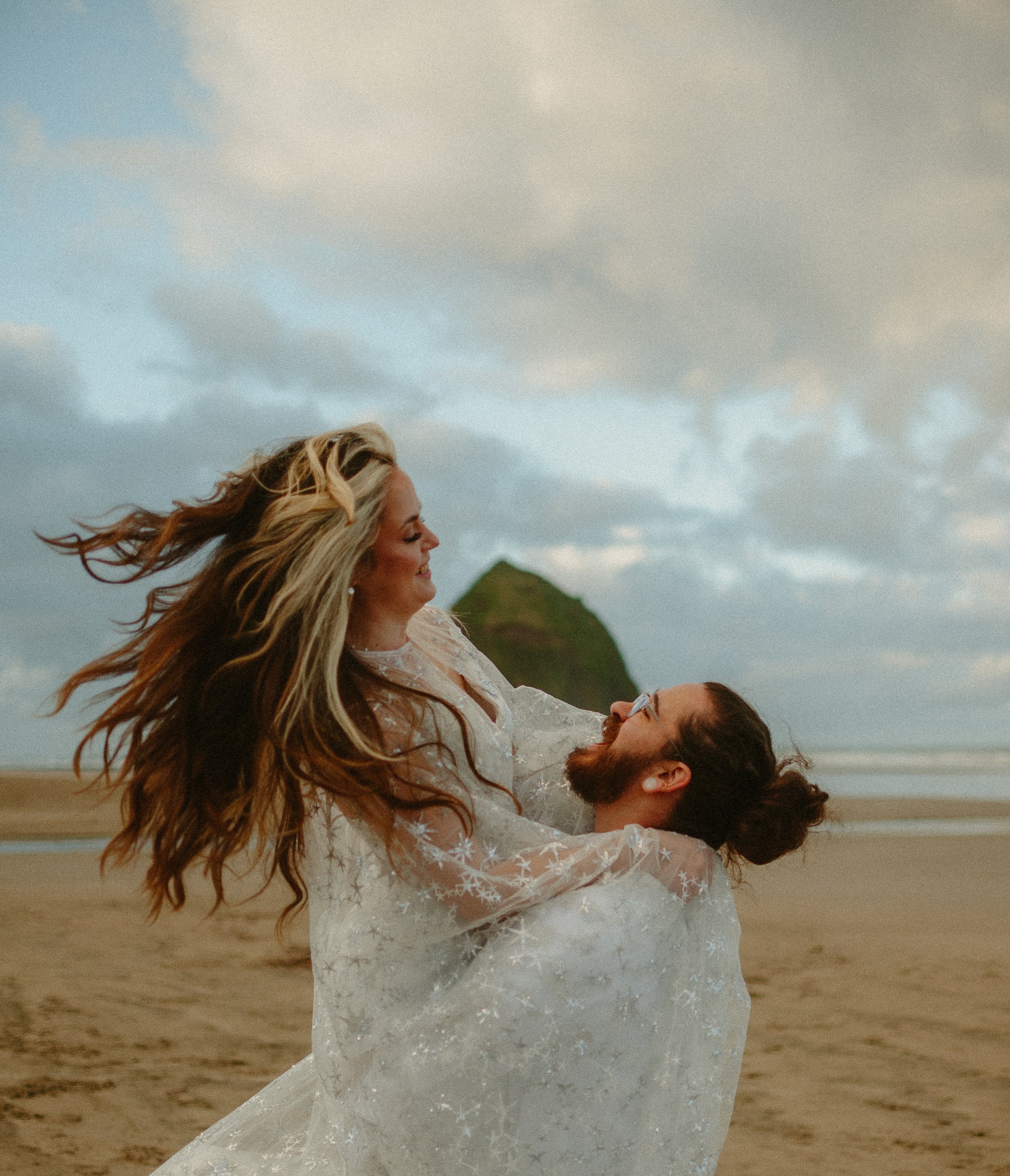 A couple in wedding attire enjoying a joyful moment on a beach with a mountain in the background.