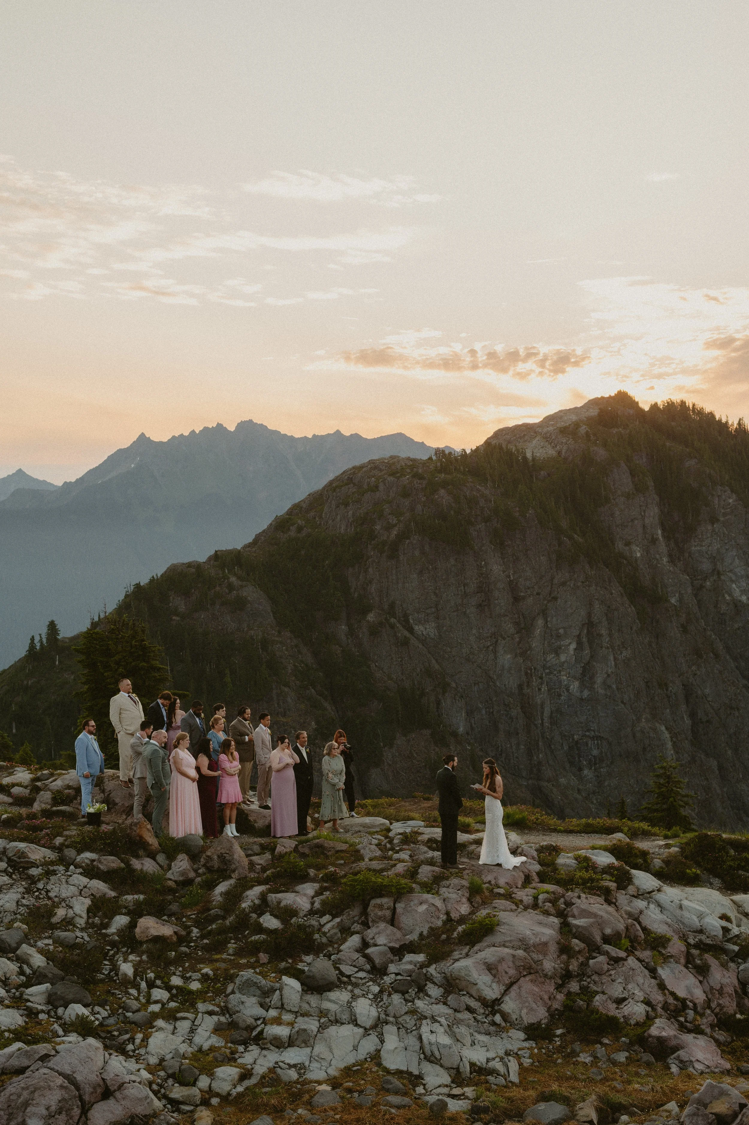 A wedding ceremony taking place outdoors on rocky terrain in a mountainous area during sunset, with a bride and groom standing before their guests under a cloudy sky.