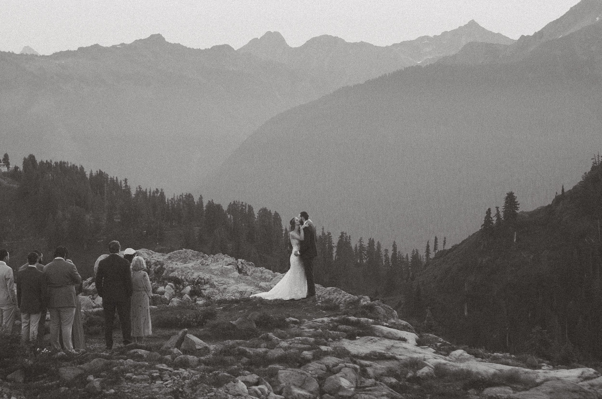 A black and white photograph of a wedding ceremony taking place outdoors in a mountainous area. The bride and groom are kissing, surrounded by seated guests, with mountains and trees in the background.