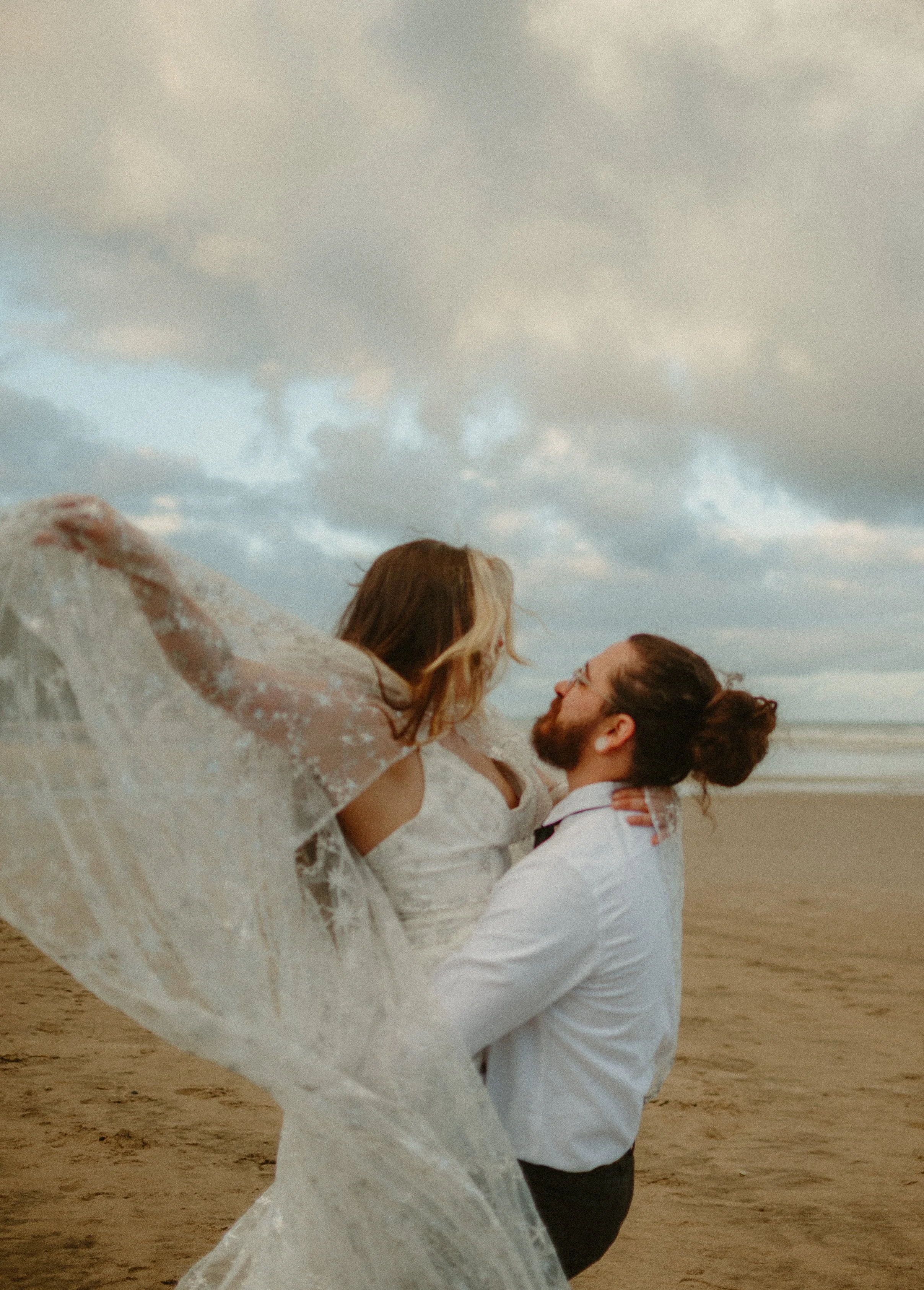 A couple on the beach, the man with a bun and glasses lifts the woman in a lace dress with a train. They are looking at each other under a cloudy sky.