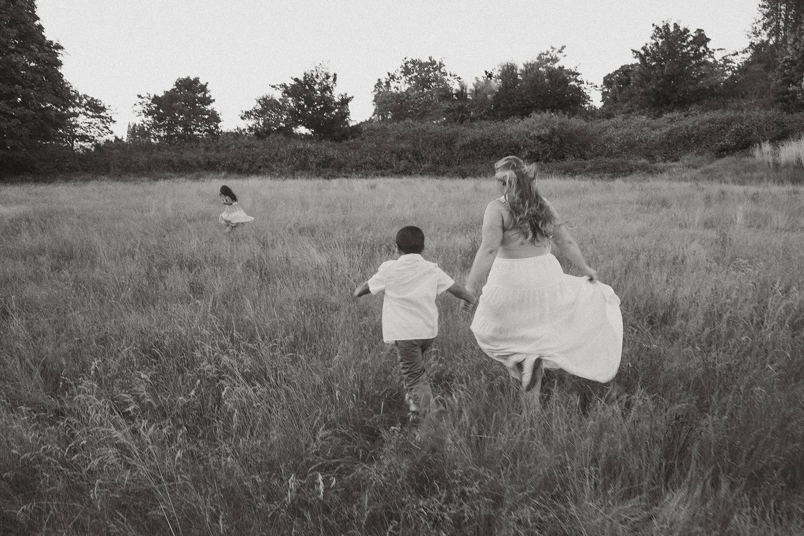 A woman holding hands with a young boy, running through a grassy field, with a girl in a dress running ahead in the distance, in black and white.