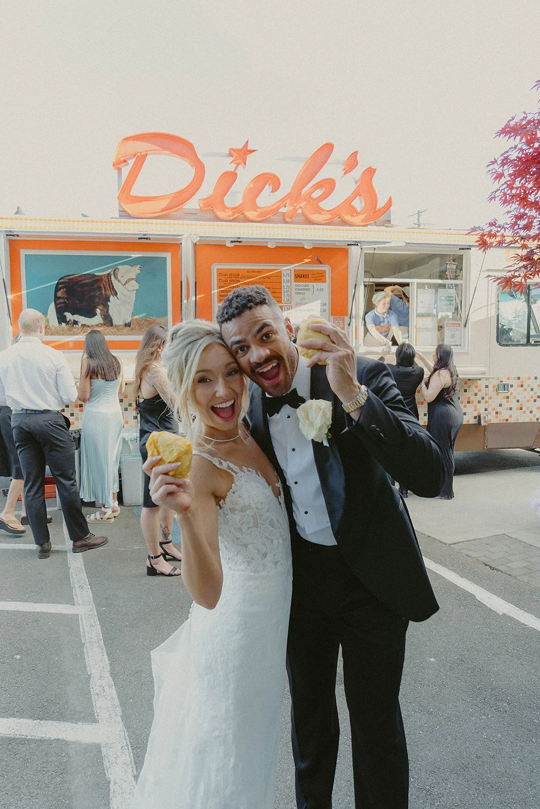 A newlywed couple in formal wedding attire enjoying hot dogs outside a vintage-style food truck called Dicks with other people waiting in line.