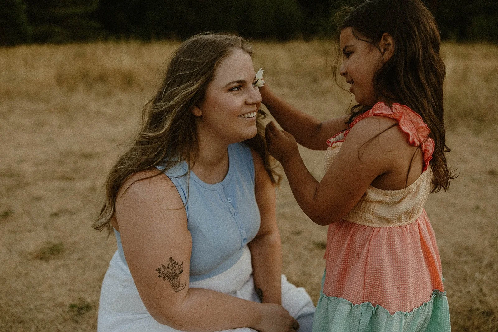 A woman with long blonde hair sitting in a field, smiling at a young girl with dark hair who is touching her face. The girl is wearing a colorful dress with ruffled sleeves.
