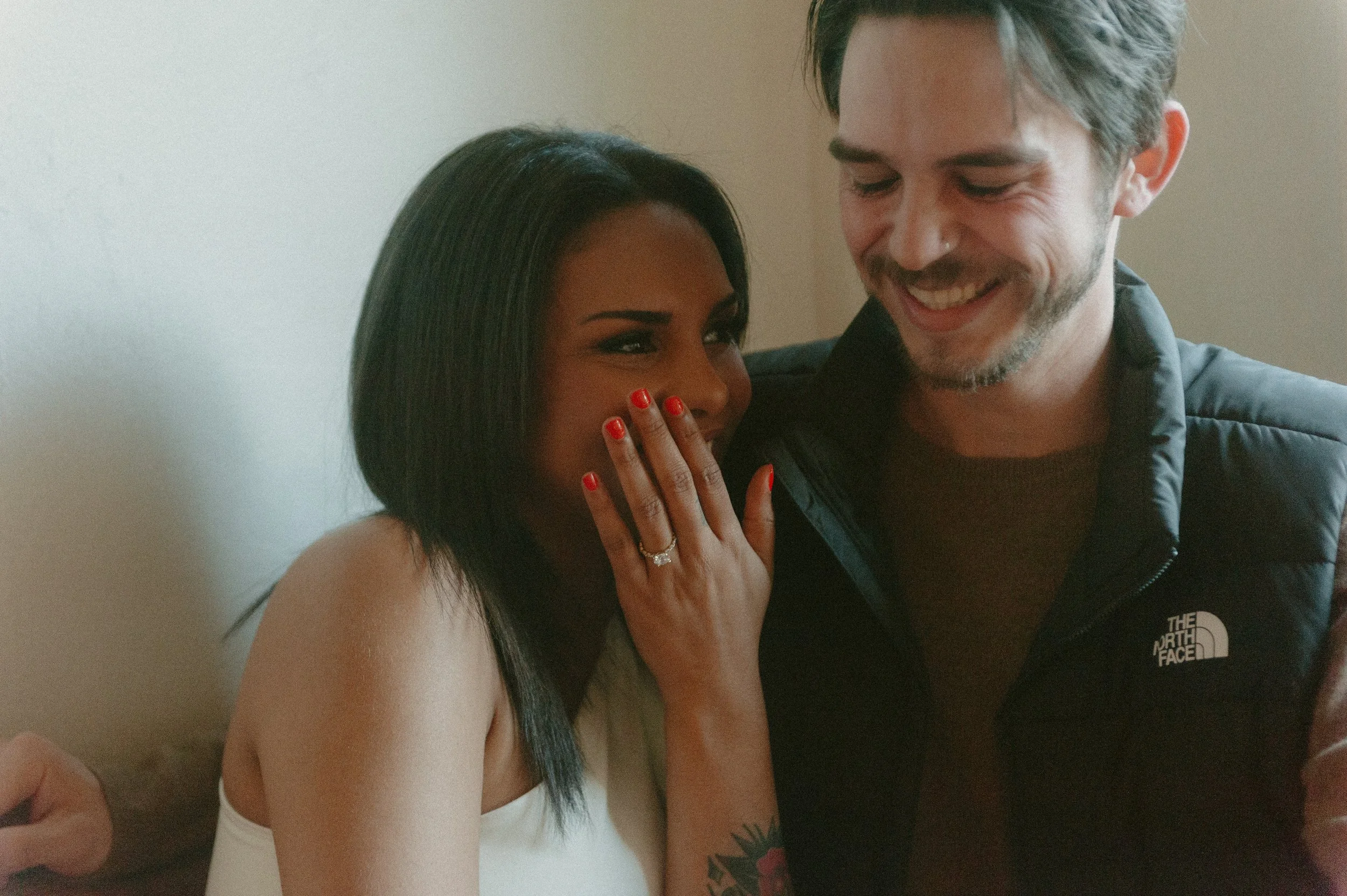 A couple shares a happy moment together indoors. The woman has medium skin tone, black hair, and is wearing a white top with red nail polish and a ring on her finger. The man has light skin, gray hair, and is wearing a dark jacket with "The North Fac