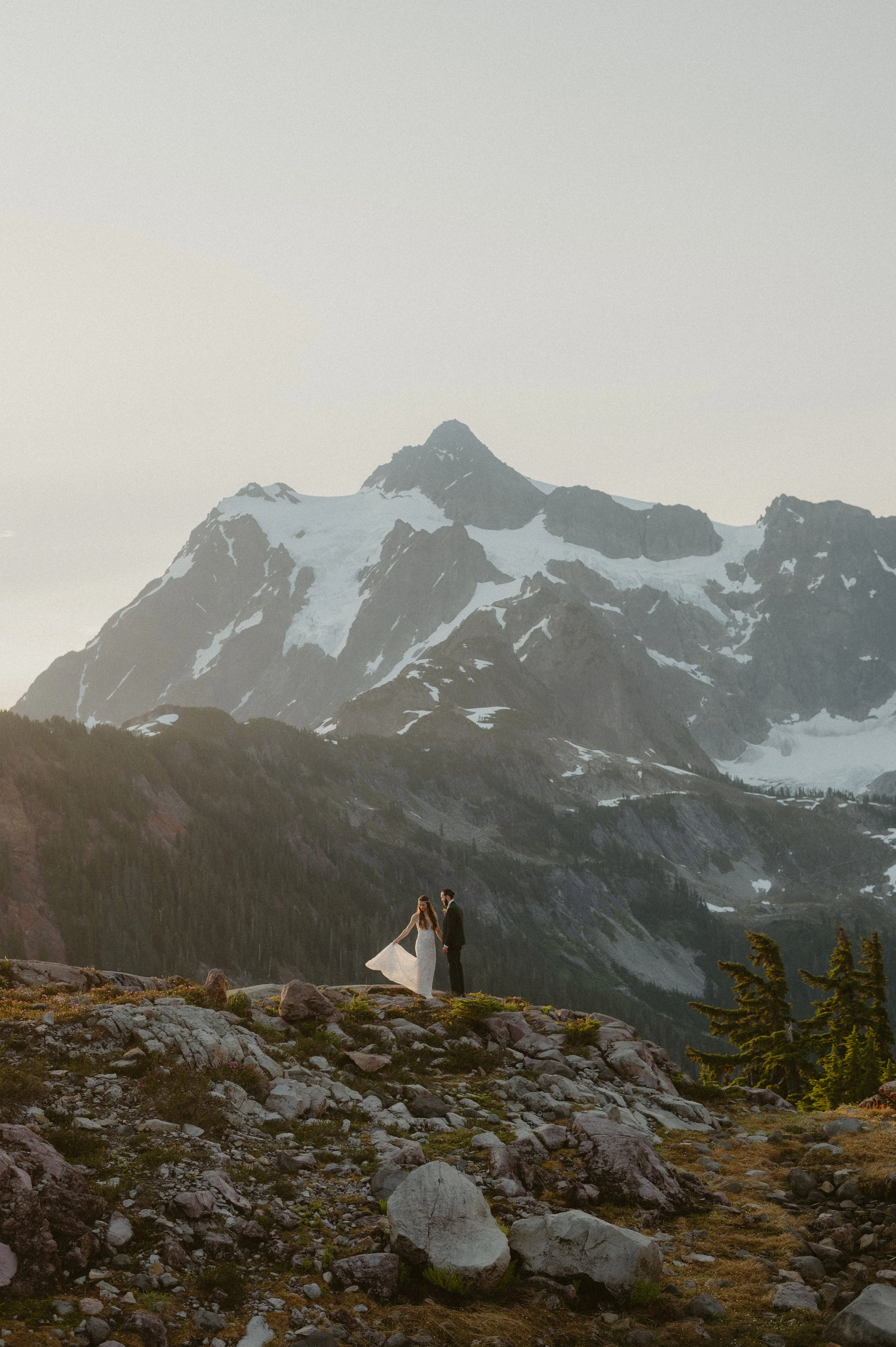 A couple dressed in wedding attire holding hands on a rocky ridge with a mountain range in the background.
