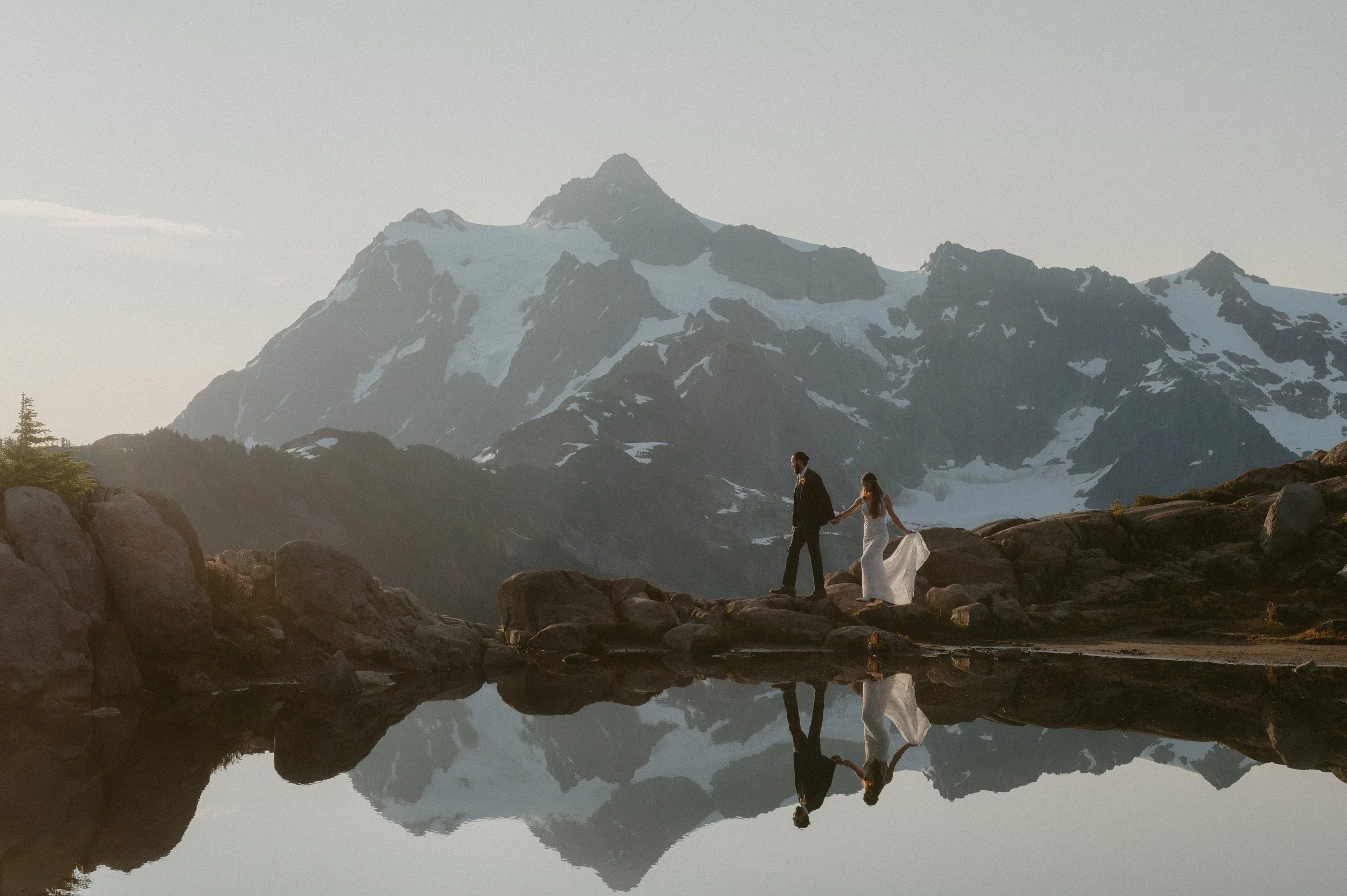 A couple in wedding attire walking hand-in-hand by a mountain lake, with snowy peaks in the background and their reflection visible in the water.