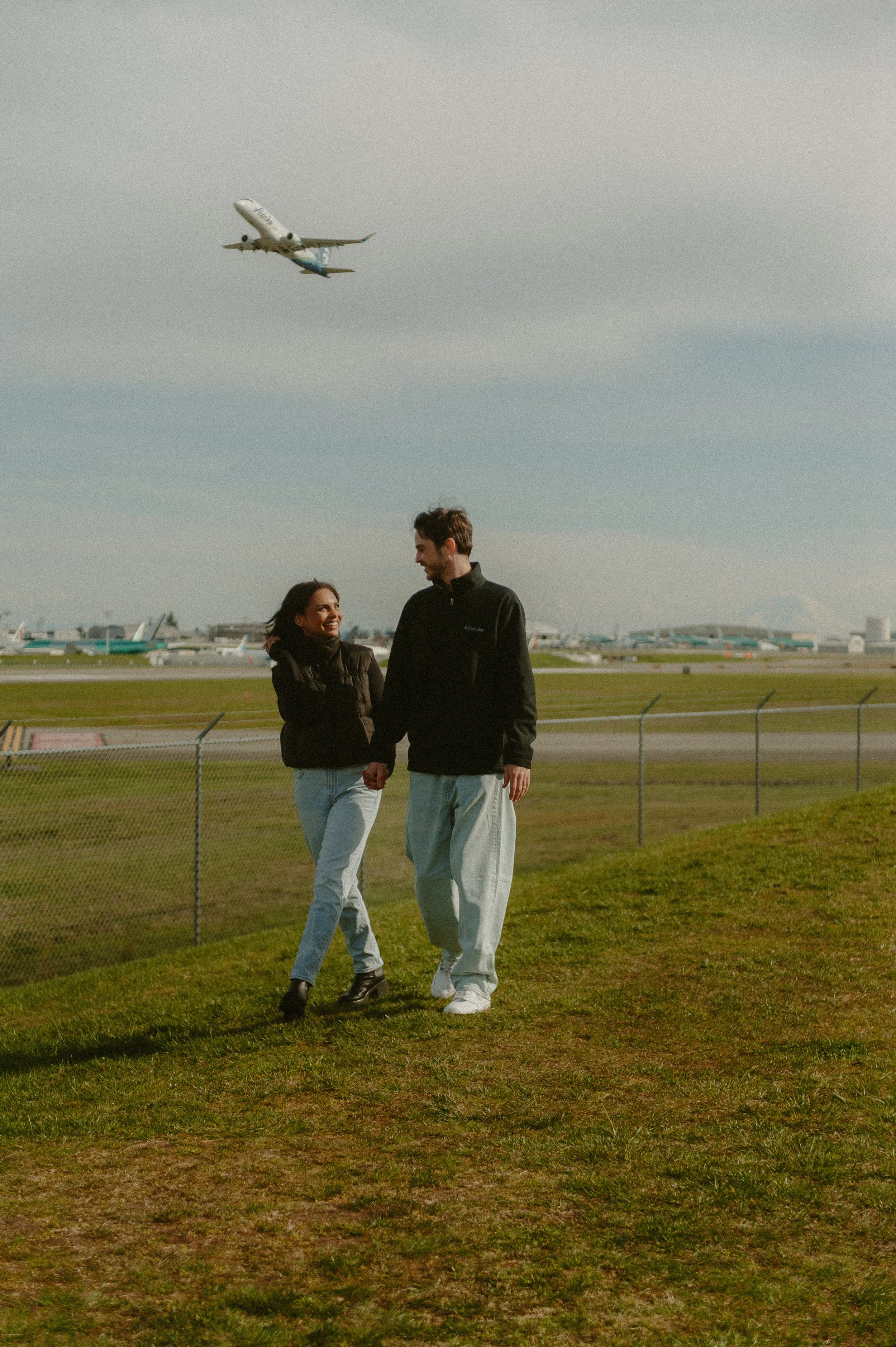 A couple walking hand in hand on a grassy area at an airport, with an airplane taking off in the background.