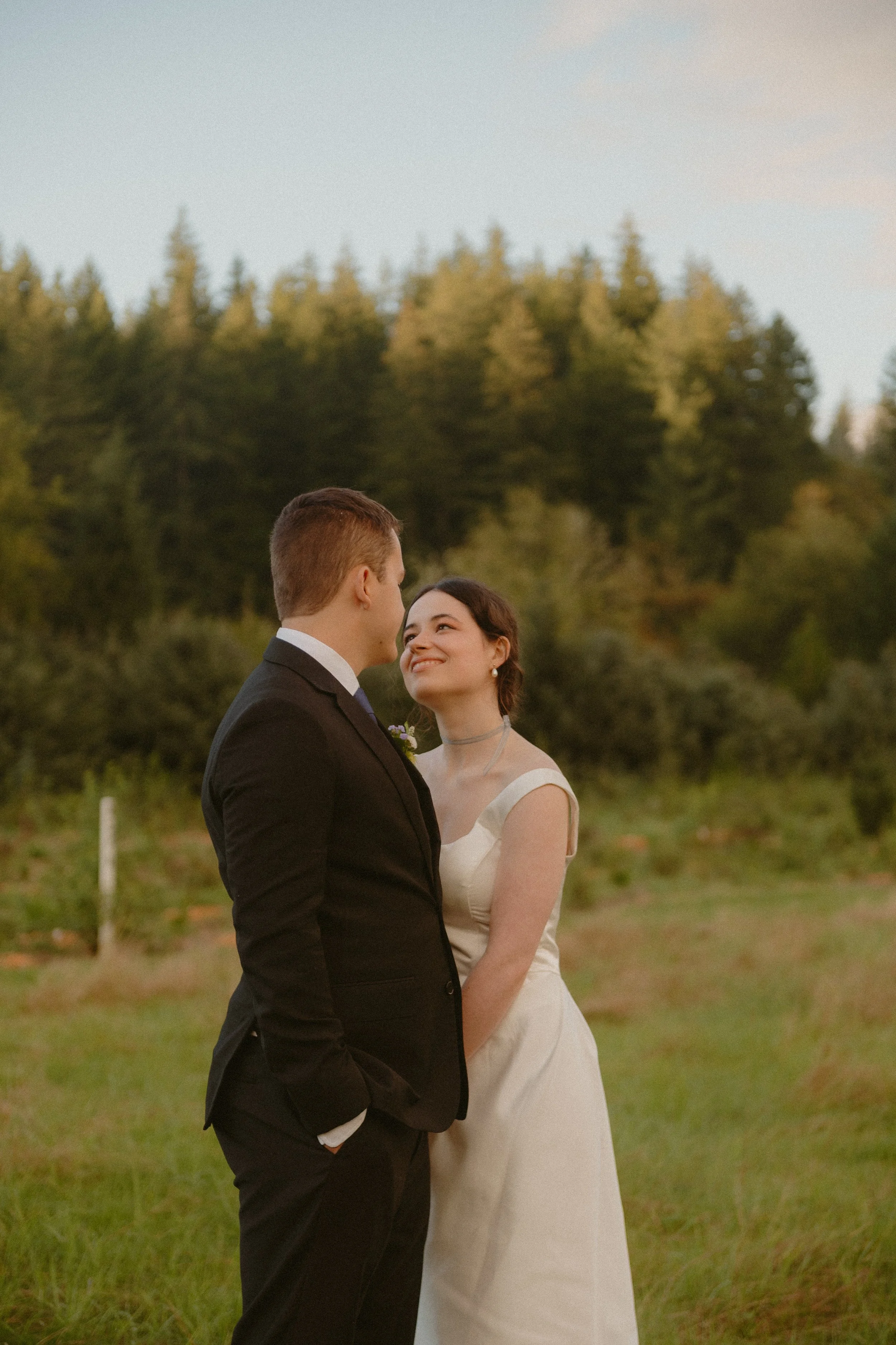 A bride and groom standing outdoors in a field, gazing at each other with smiles, against a backdrop of trees during sunset.