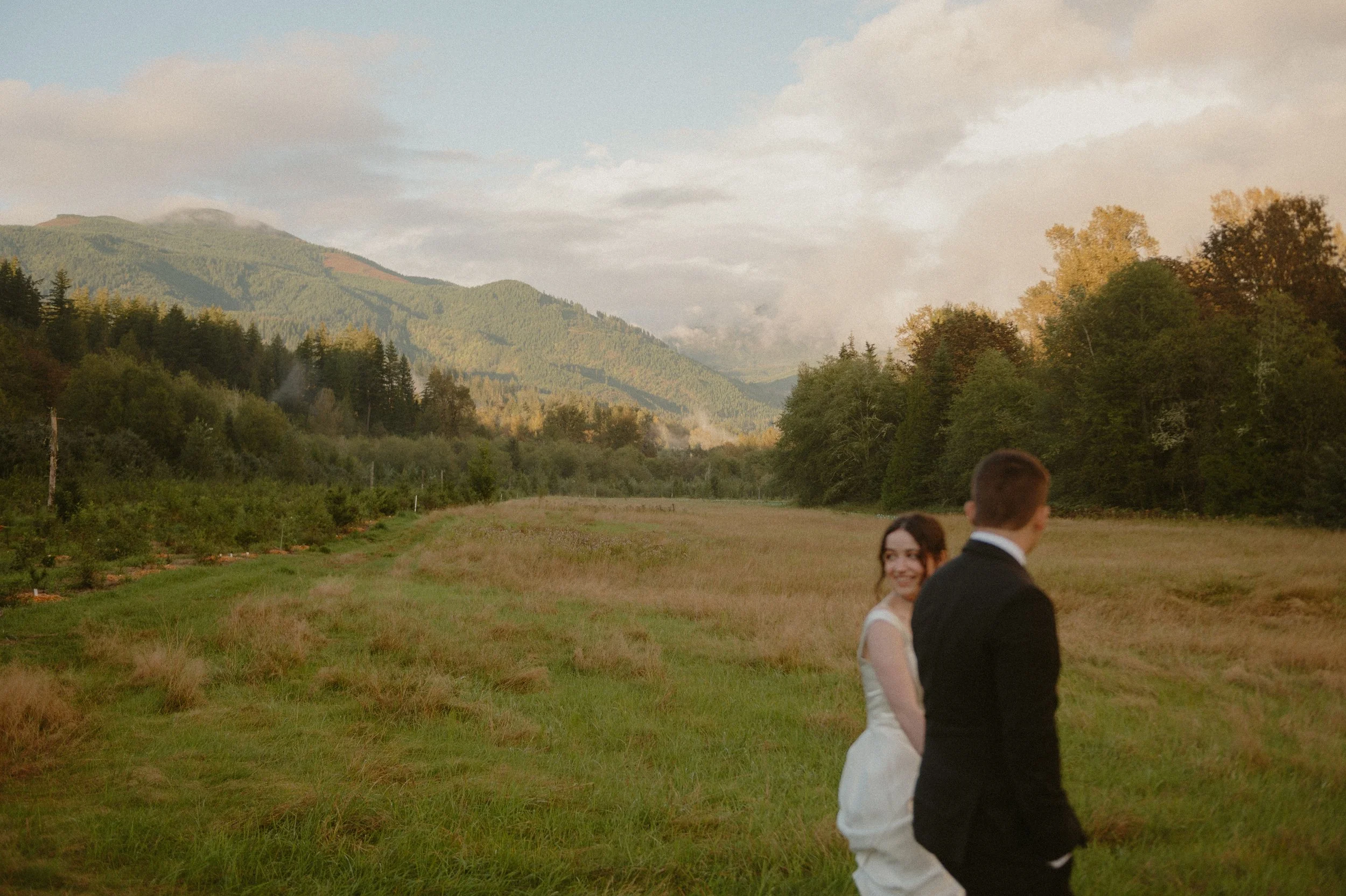 A couple dressed in wedding attire walking through a grassy field with mountains and trees in the background, smiling at each other.