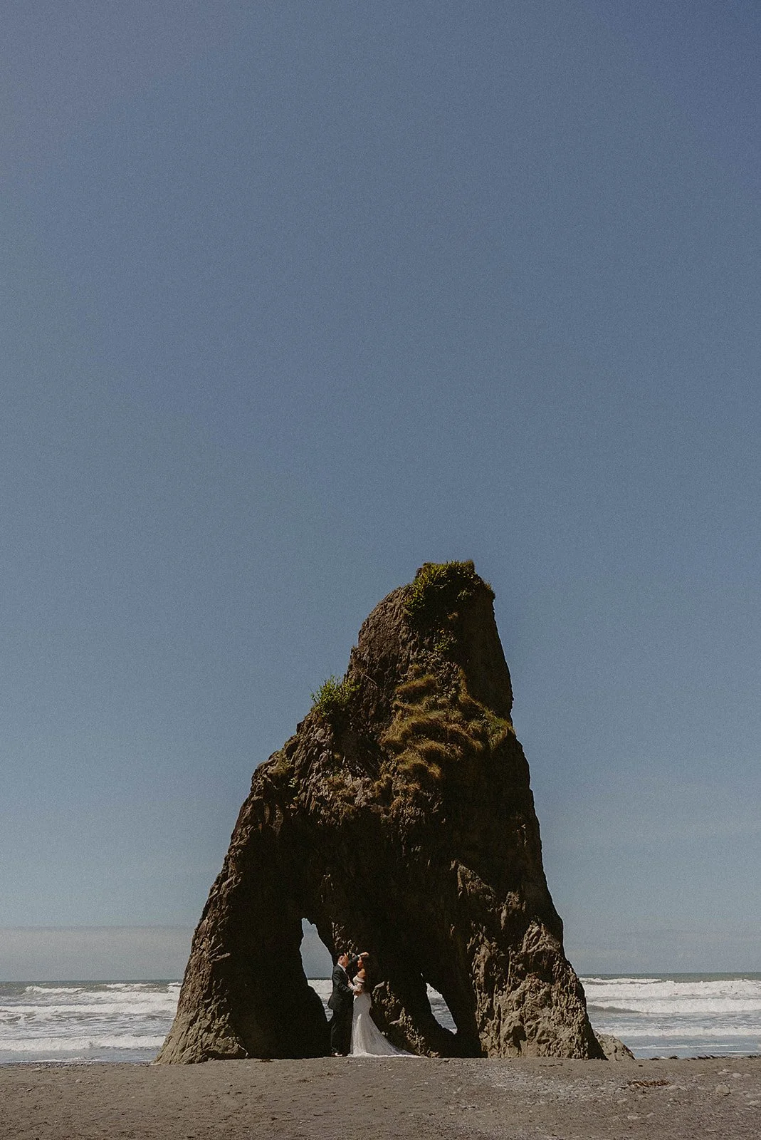 A couple dressed in wedding attire standing underneath a large sea arch on a beach, with the ocean and a clear blue sky in the background.