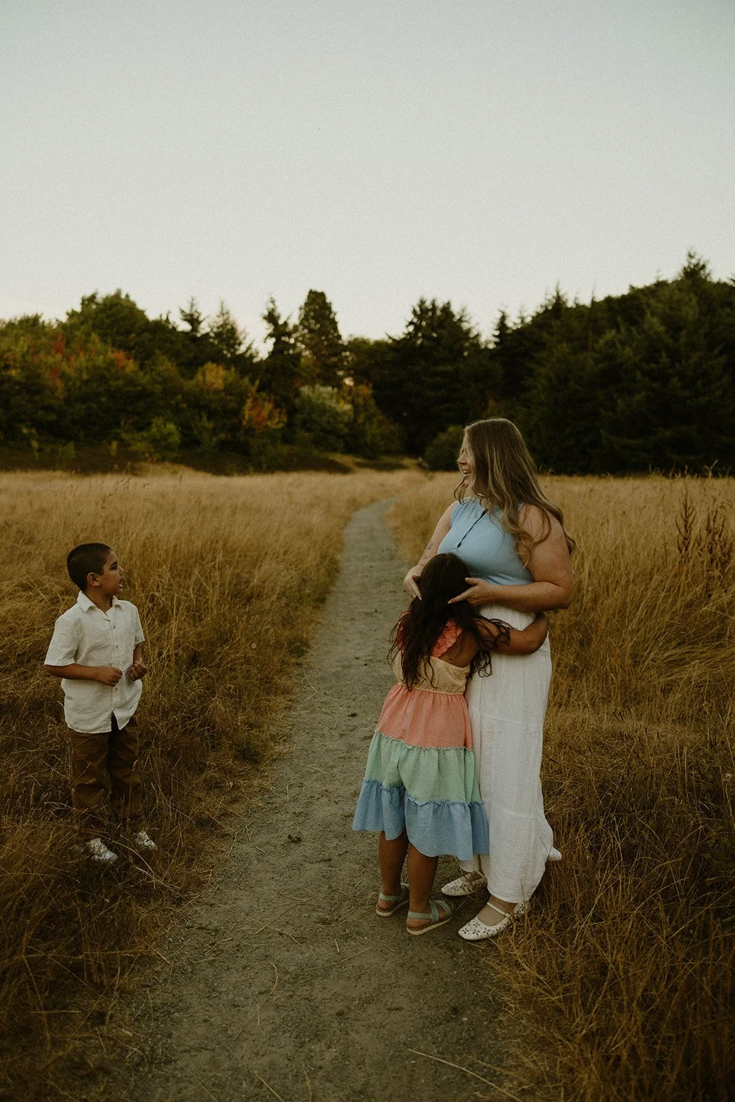 A woman hugging two children in a field with a dirt path, while a boy stands nearby looking on, during sunset.