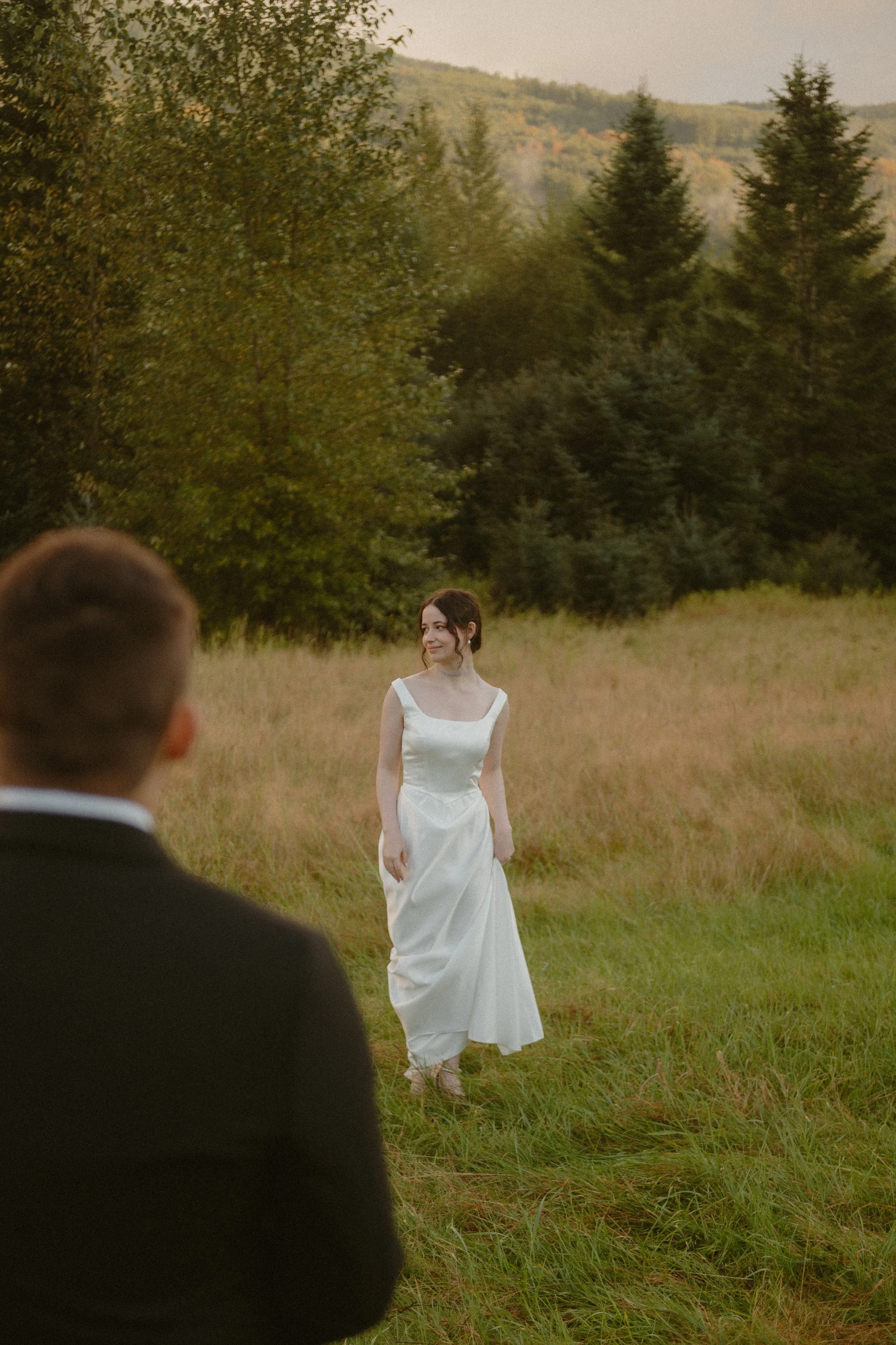 A bride in a white wedding dress walking on a grassy field with trees and hills in the background, while a groom in a suit watches her.