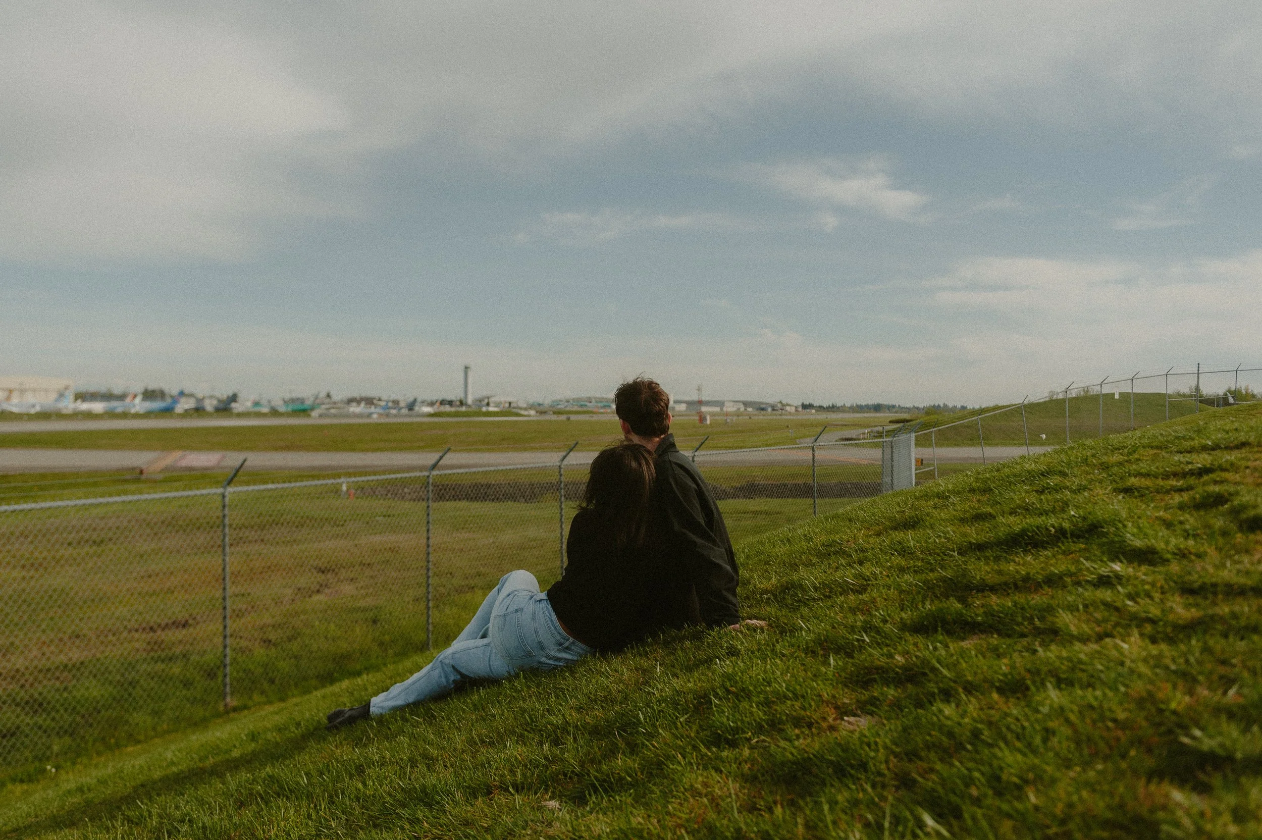 A man and woman sitting on a grassy hill near a fence at an airport, watching airplanes on the runway under a partly cloudy sky.