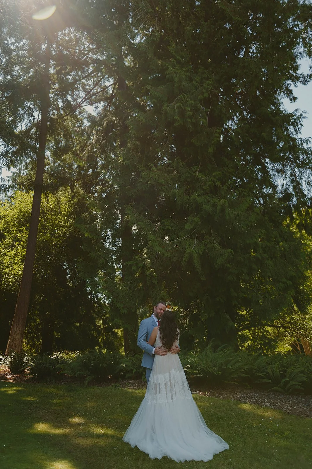A bride and groom embrace outdoors in a lush green garden under tall trees, sunlight filtering through the leaves.