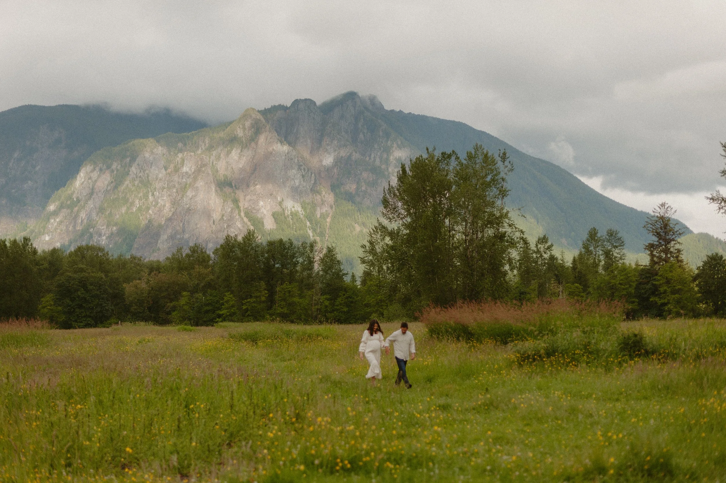 A couple walks hand in hand through a grassy field with yellow flowers, with mountains and trees in the background under cloudy skies.